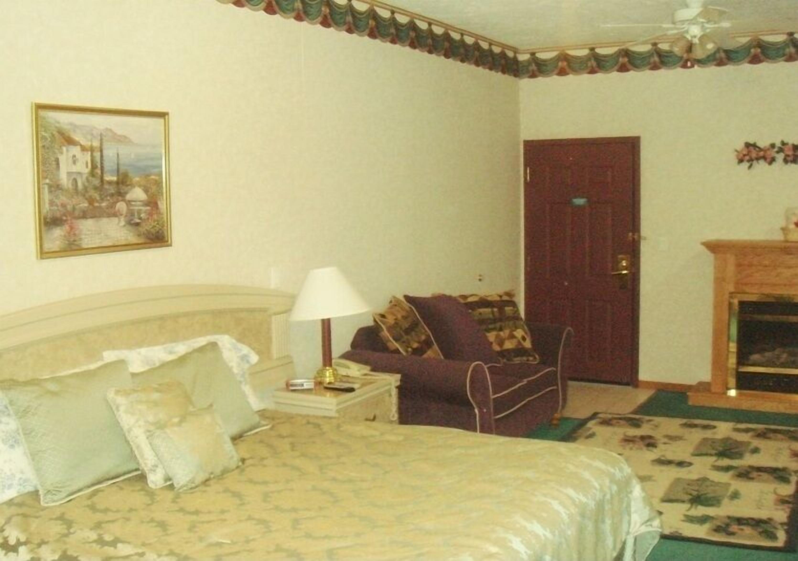 Um quarto com cama, mesa de cabeceira, abajur, poltrona, porta e lareira.