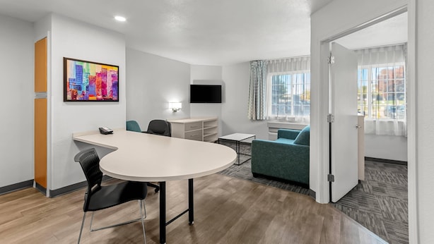 A modern hotel room with a round table, a sofa, a TV mounted on the wall, and a window with curtains. - MainStay Suites Louisville Jeffersontown (Louisville)