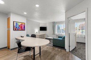 Una habitación de hotel moderna con una mesa redonda, un sofá, un televisor montado en la pared y un ventanal con cortinas.