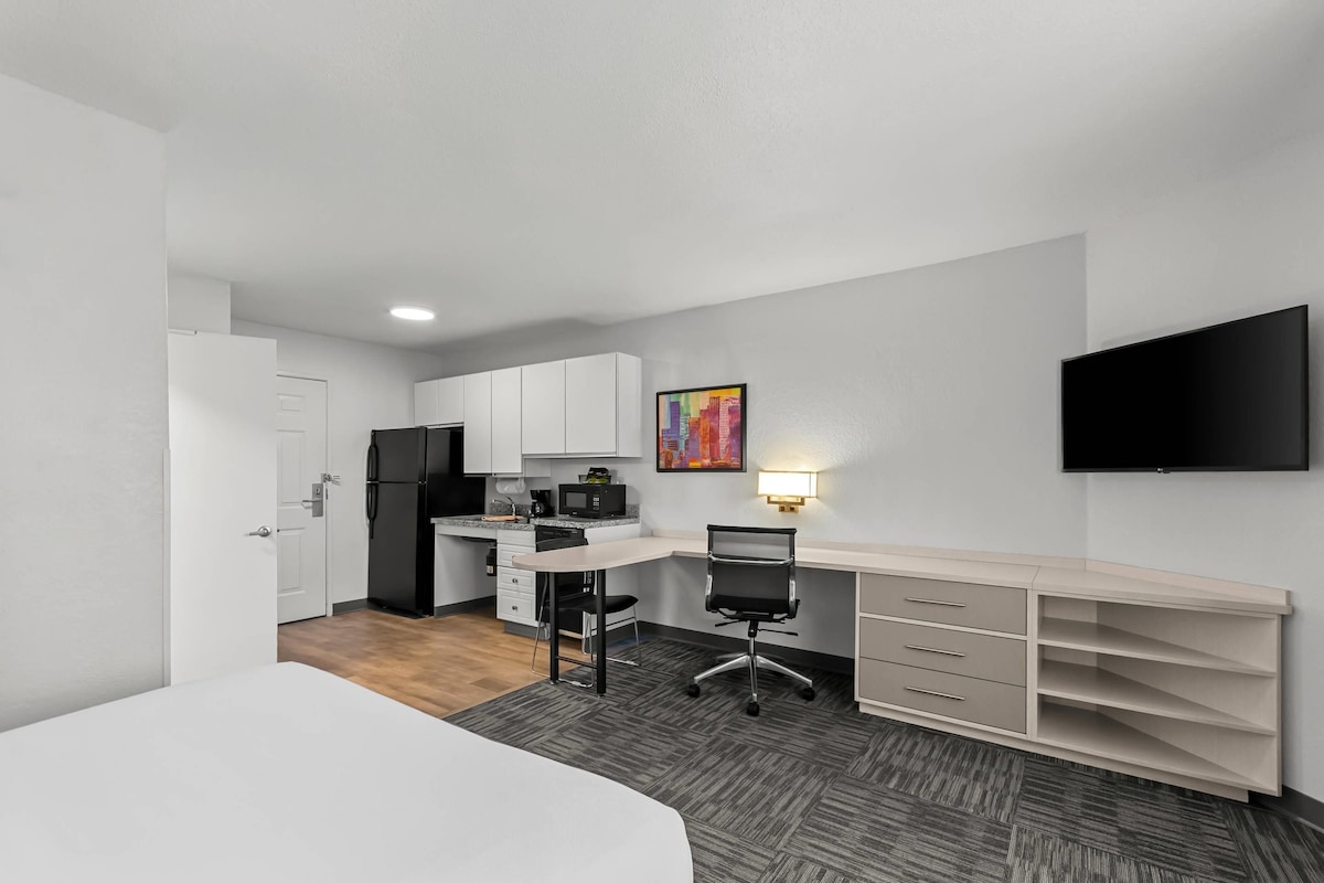 A compact hotel room with a kitchenette, a bed, a desk with a chair, and a TV mounted on the wall.