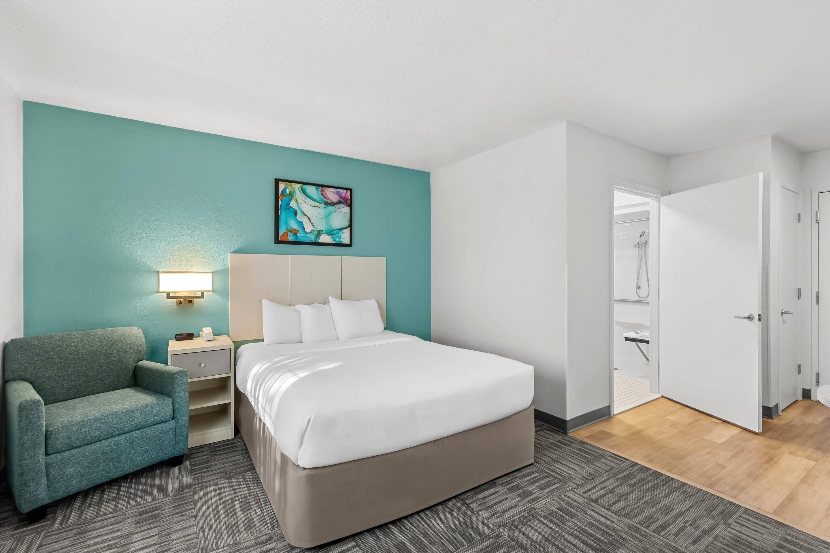 Una habitación de hotel con cama, sillón, mesita de noche, un cuadro y la puerta del baño.