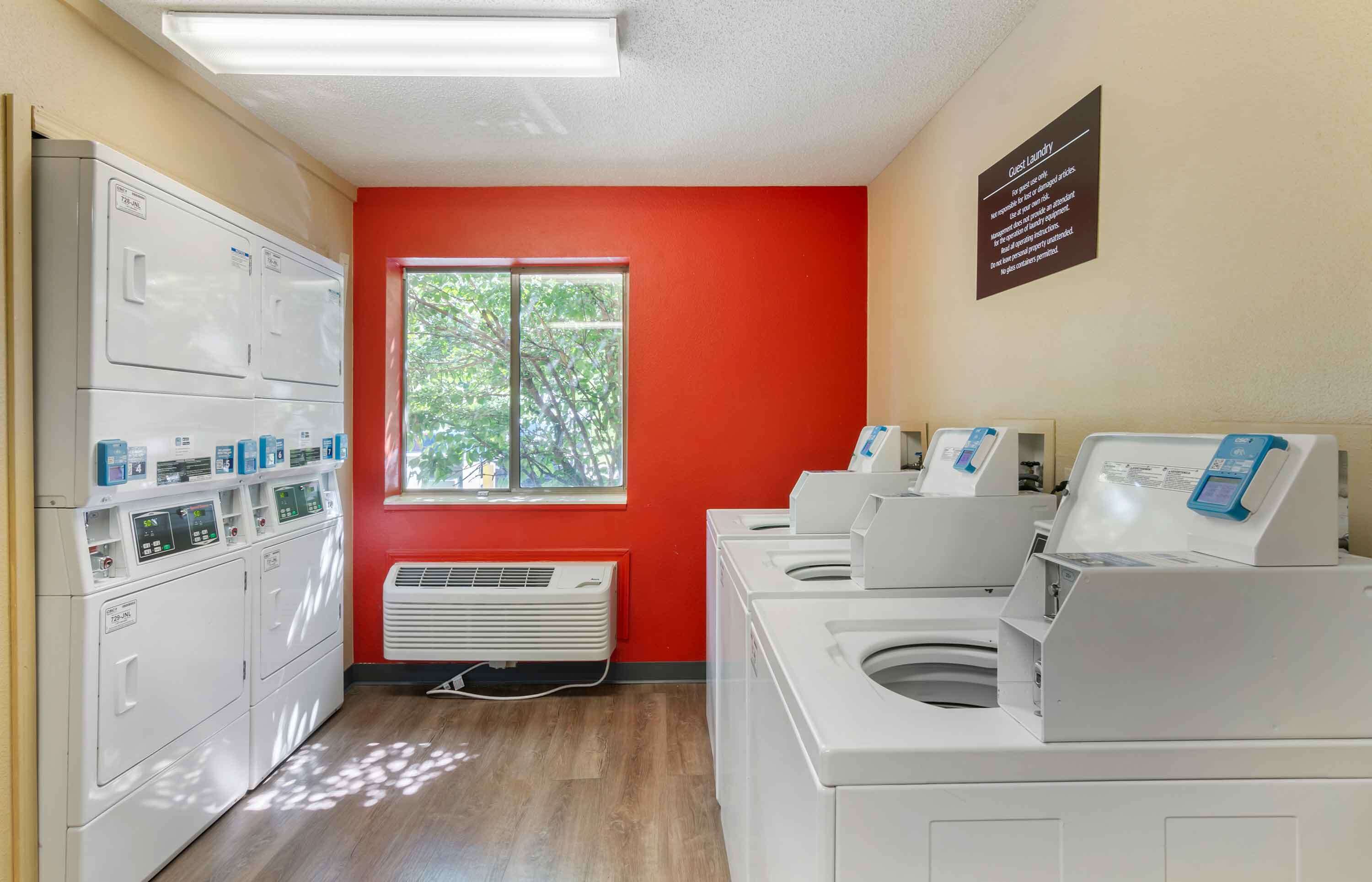 Une buanderie équipée de plusieurs machines à laver et de plusieurs sèche-linge, une fenêtre donnant sur des arbres et un panneau affiché sur le mur.
