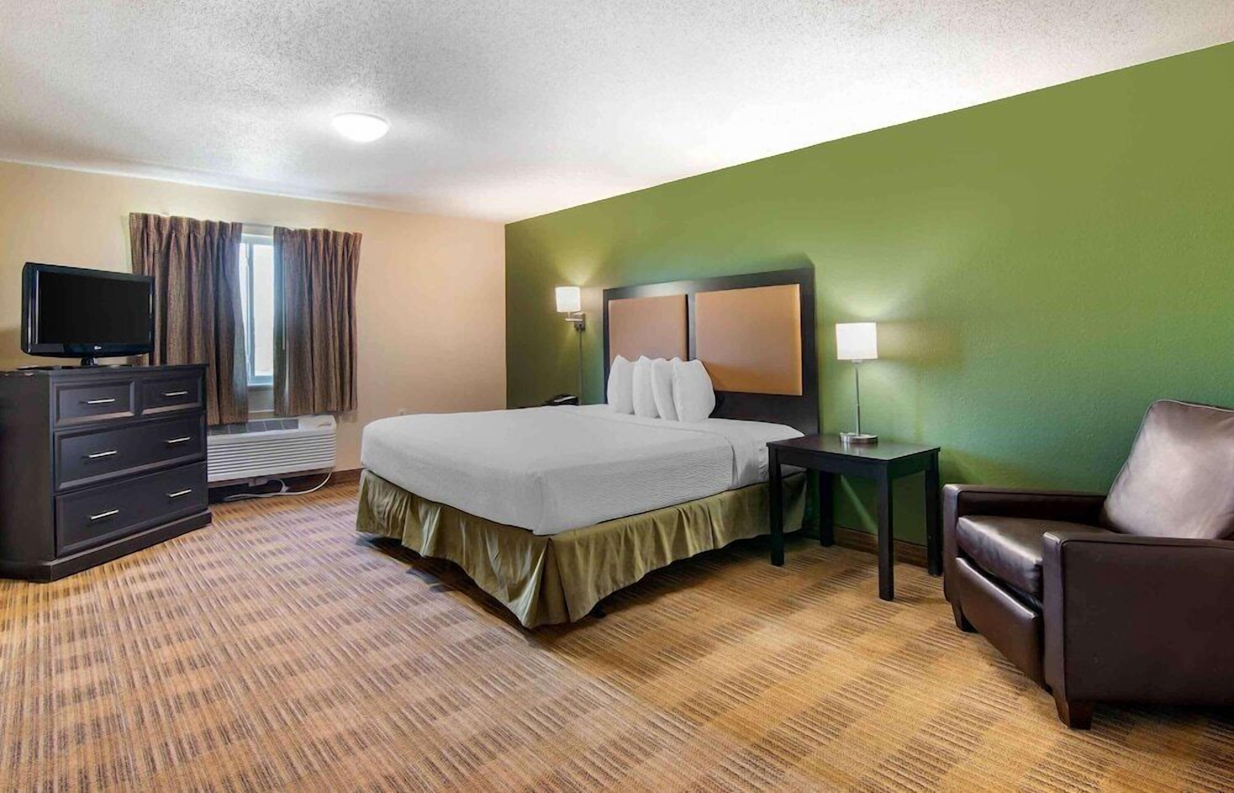 Quarto de hotel com cama, televisão, penteadeira, cadeira e abajur.