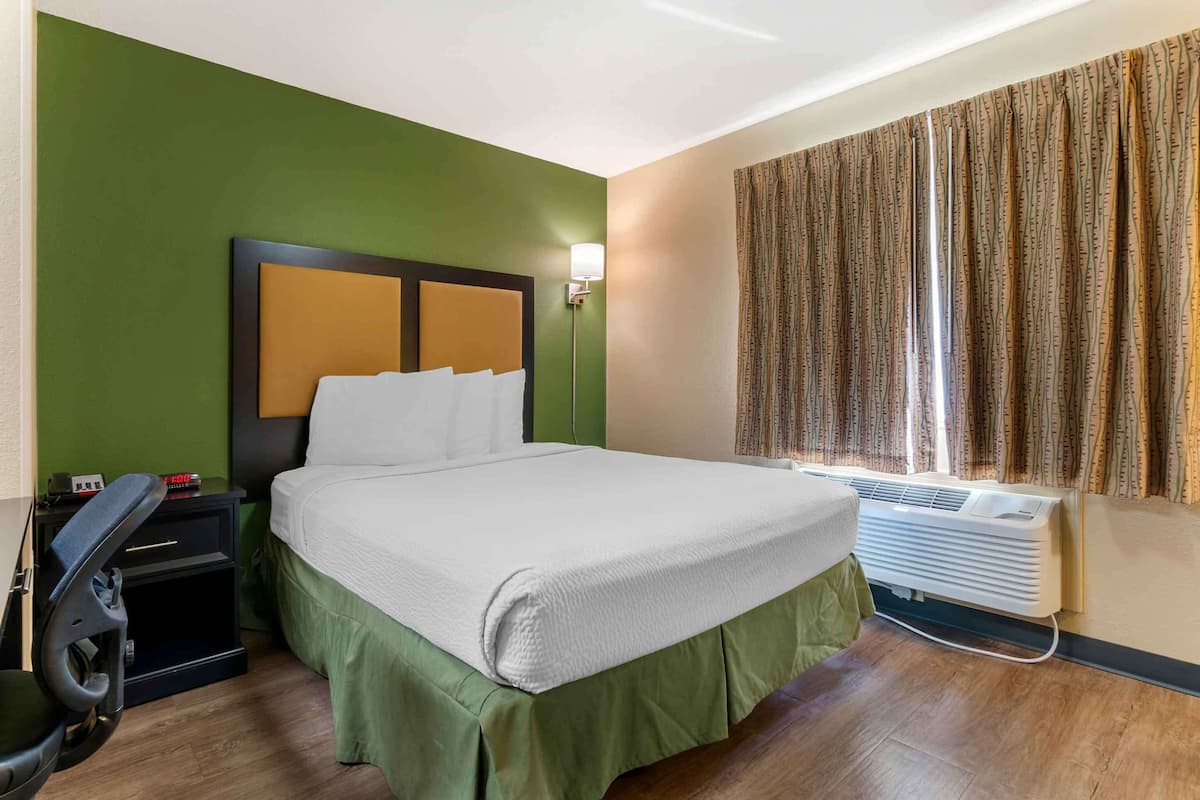 A hotel room with a bed, a desk with a chair, a standing heater, and a window with curtains.