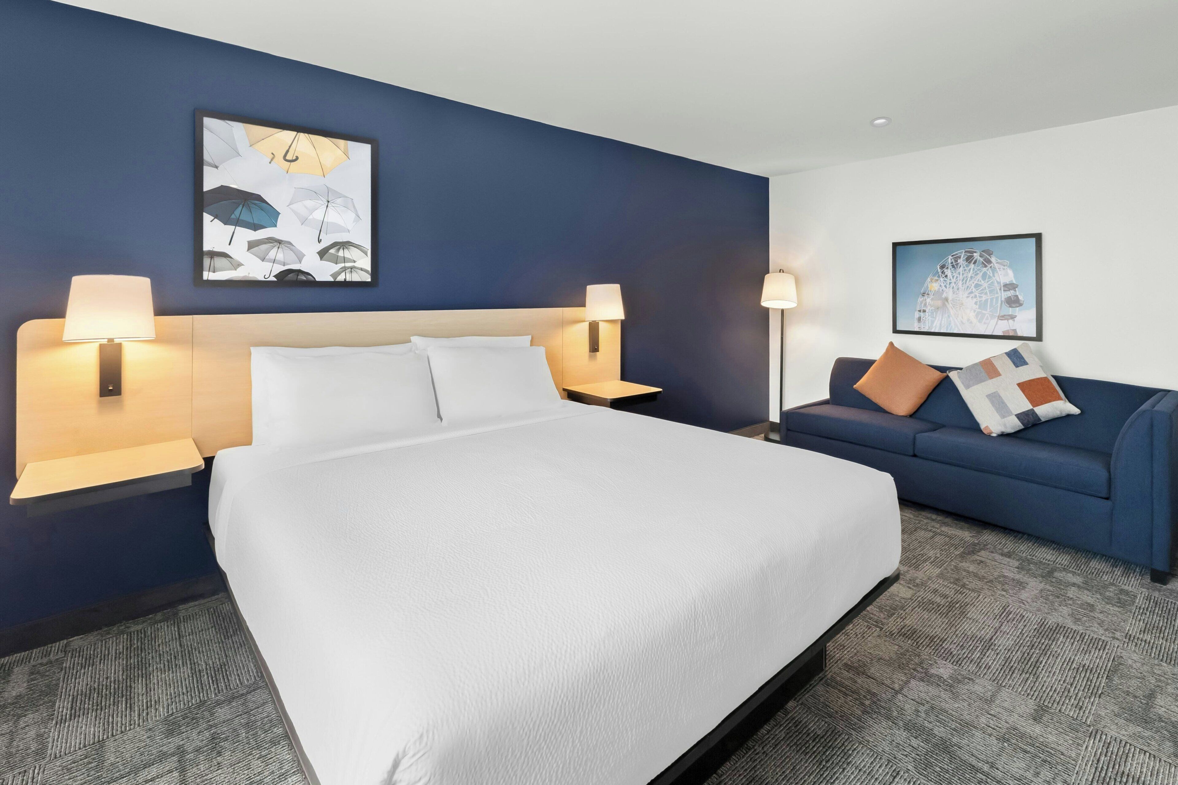 Quarto de hotel com cama, duas luminárias de cabeceira, um sofá azul com almofada e duas imagens emolduradas nas paredes.