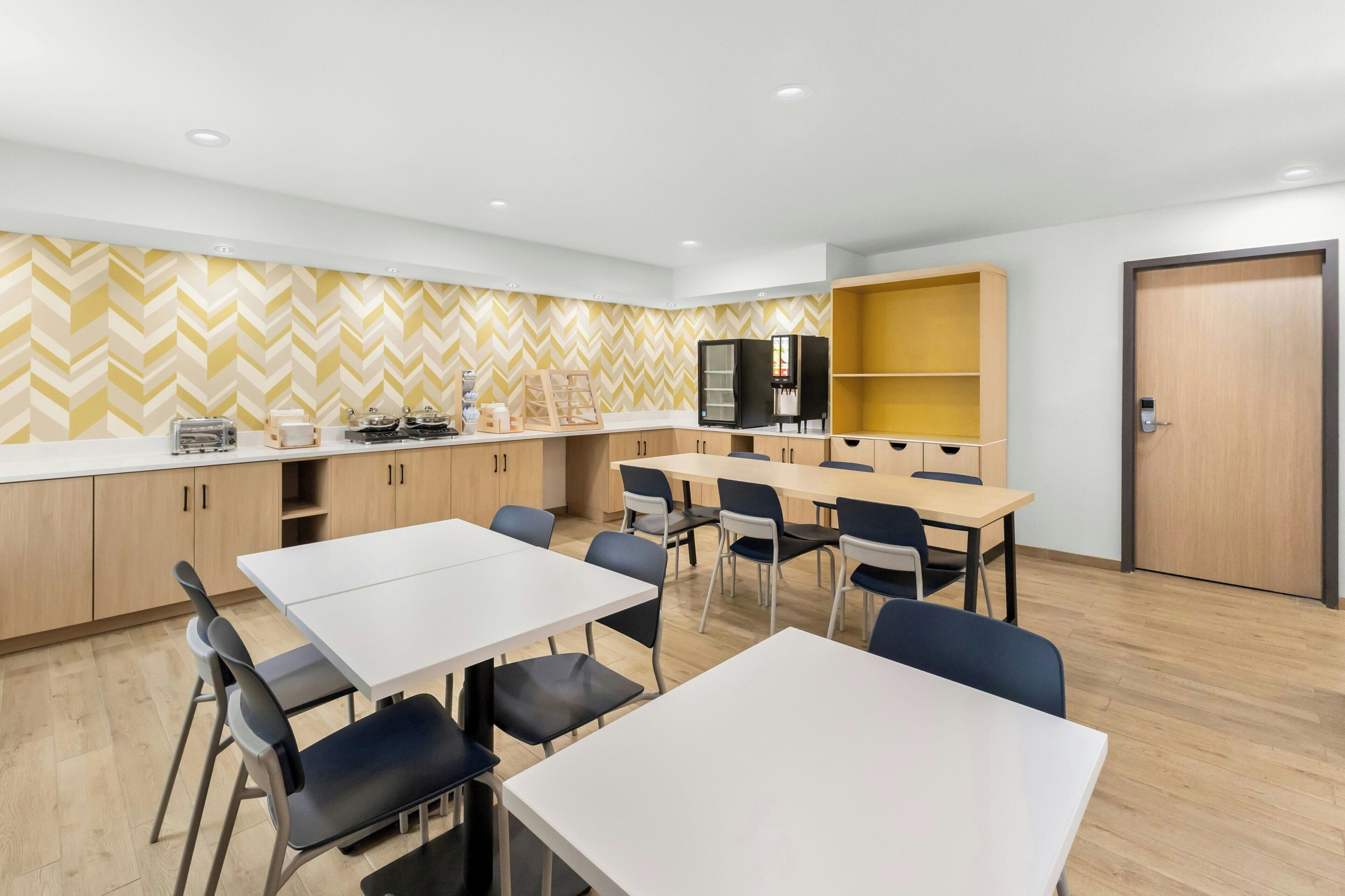 Une cuisine moderne avec des meubles blancs, un réfrigérateur noir et un four à micro-ondes. On y trouve deux tables blanches avec des chaises, et un mur jaune orné d’un motif en chevrons.