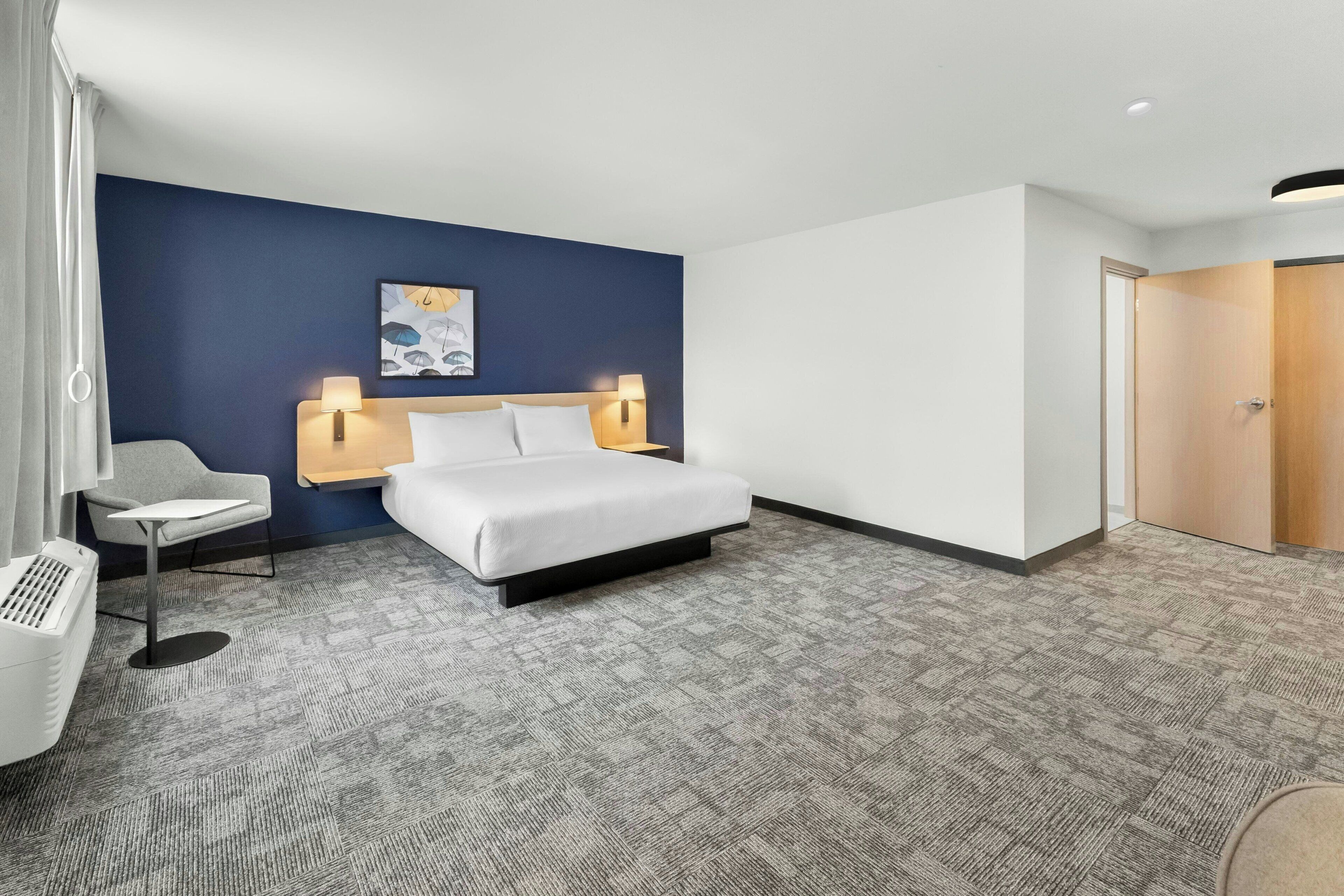 A hotel room with a bed, bedside lamps, a chair, a small table, and a wall-mounted artwork.