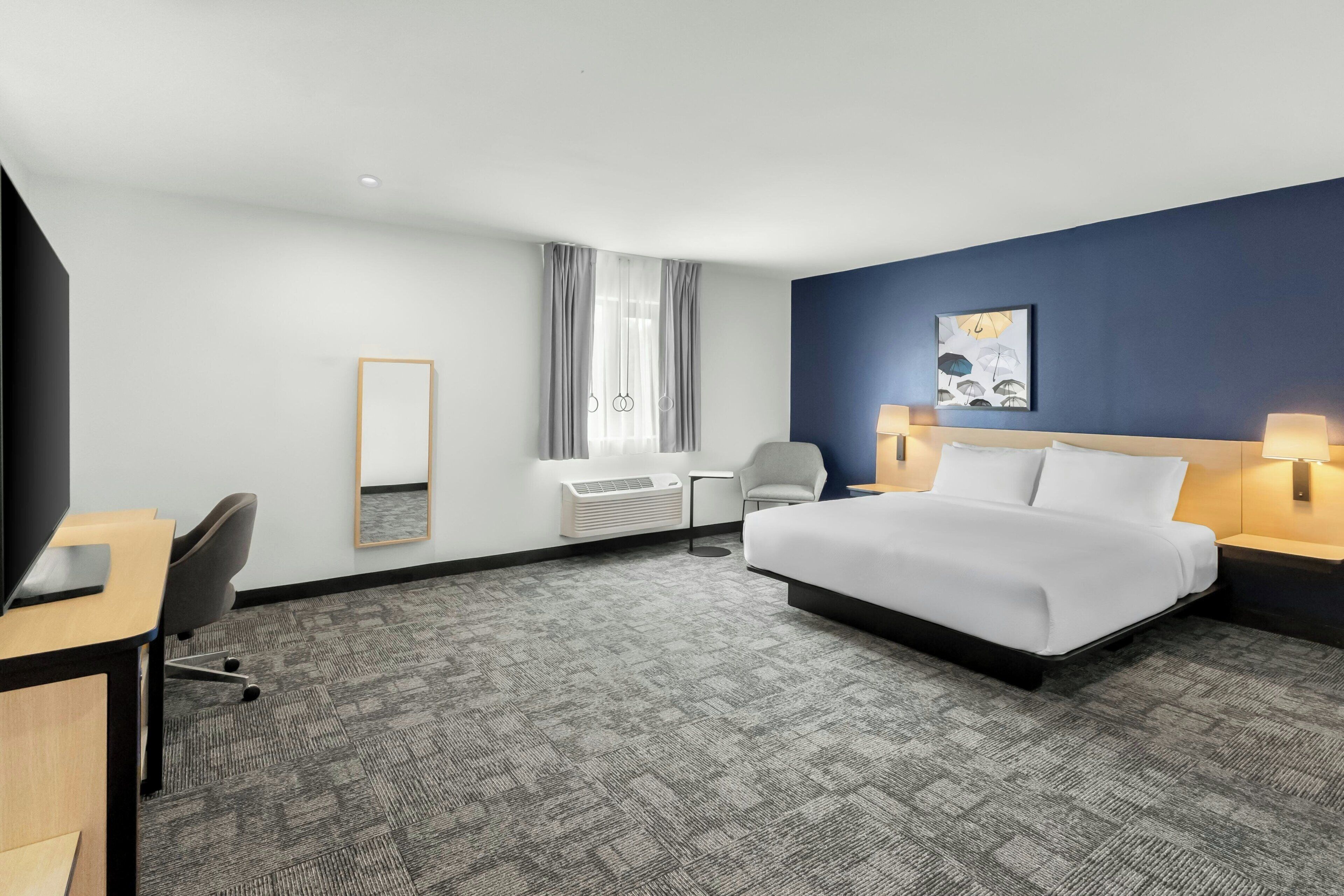 A hotel room with a large bed, a desk, a chair, a mirror, a window with curtains, and a wall-mounted artwork.