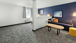 A hotel room with a sofa, a small table, a chair, a bed, a window with curtains, a wall-mounted air conditioner, and two wall art pieces.