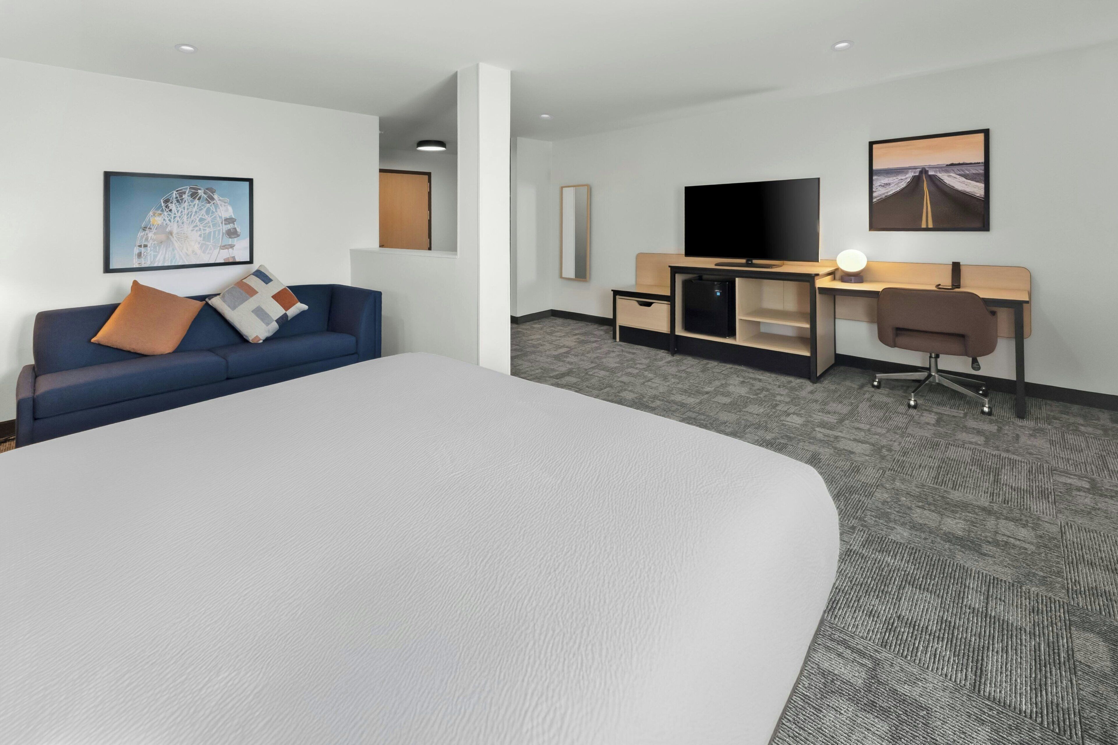 A hotel room with a large bed, a sofa, a desk with a computer, a TV, and artwork on the walls.