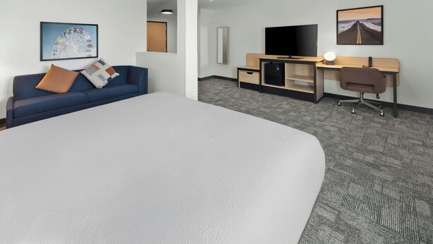 A hotel room with a large bed, a sofa, a desk with a computer, a TV, and artwork on the walls.