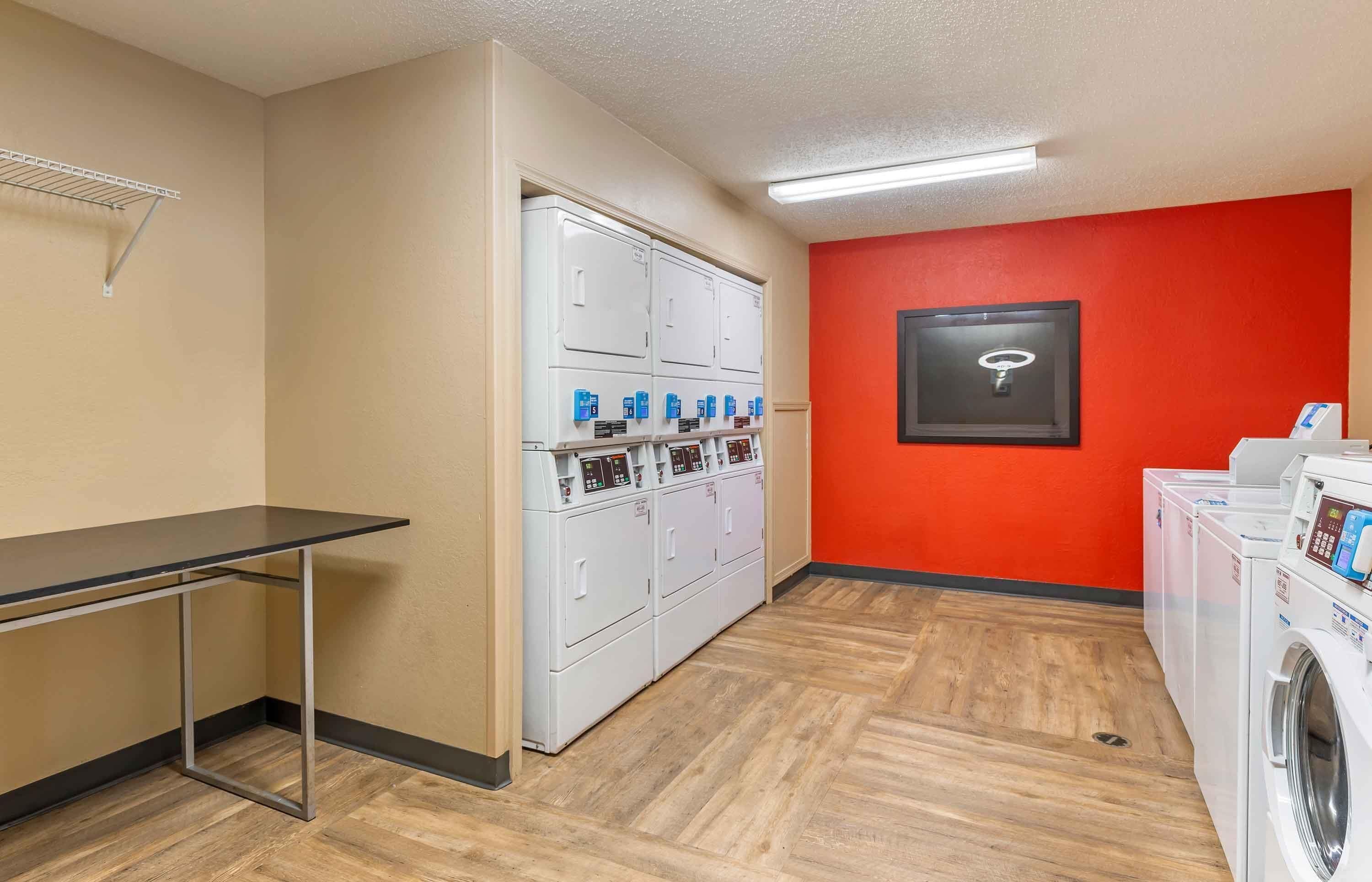 A laundry room with a metal drying rack, a row of white dryers, and a red wall with a framed picture.