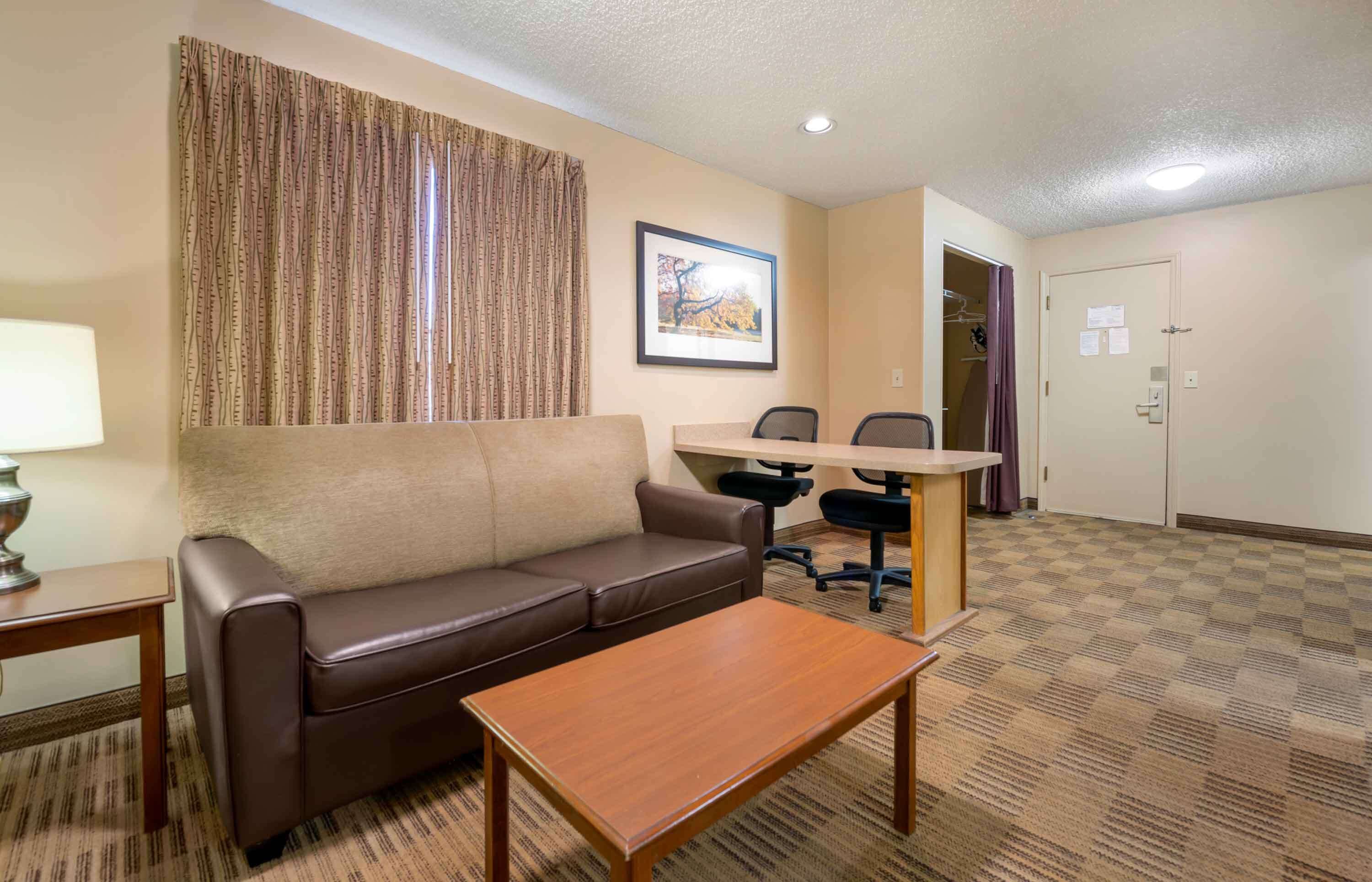 Une chambre d’hôtel avec un canapé en cuir marron, une table basse en bois, un bureau avec deux chaises, un rideau à motifs et un tableau encadré au mur.