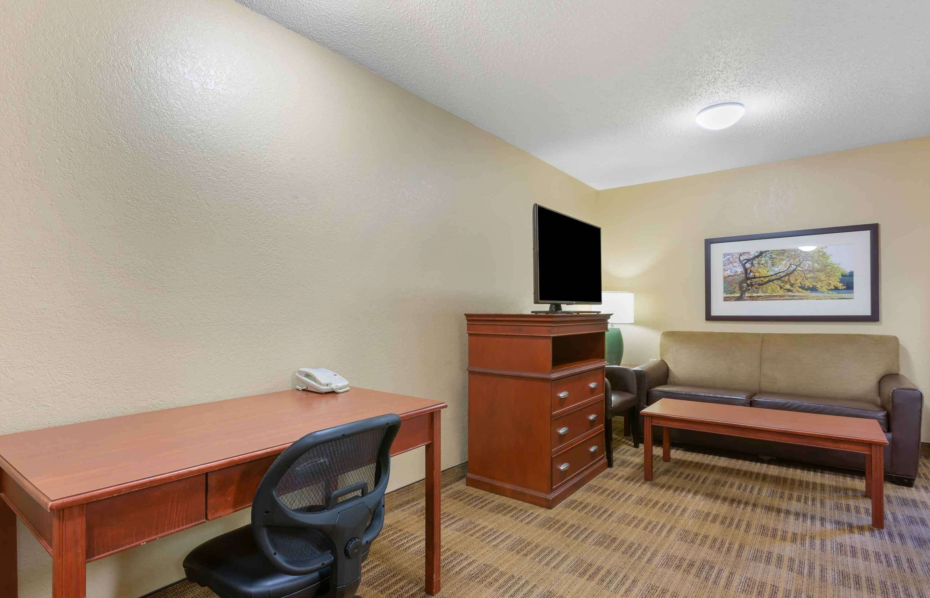 Ein Hotelzimmer mit einem Holztisch, einem Sessel, einer Couch, einem Fernseher und einem gerahmten Bild an der Wand.