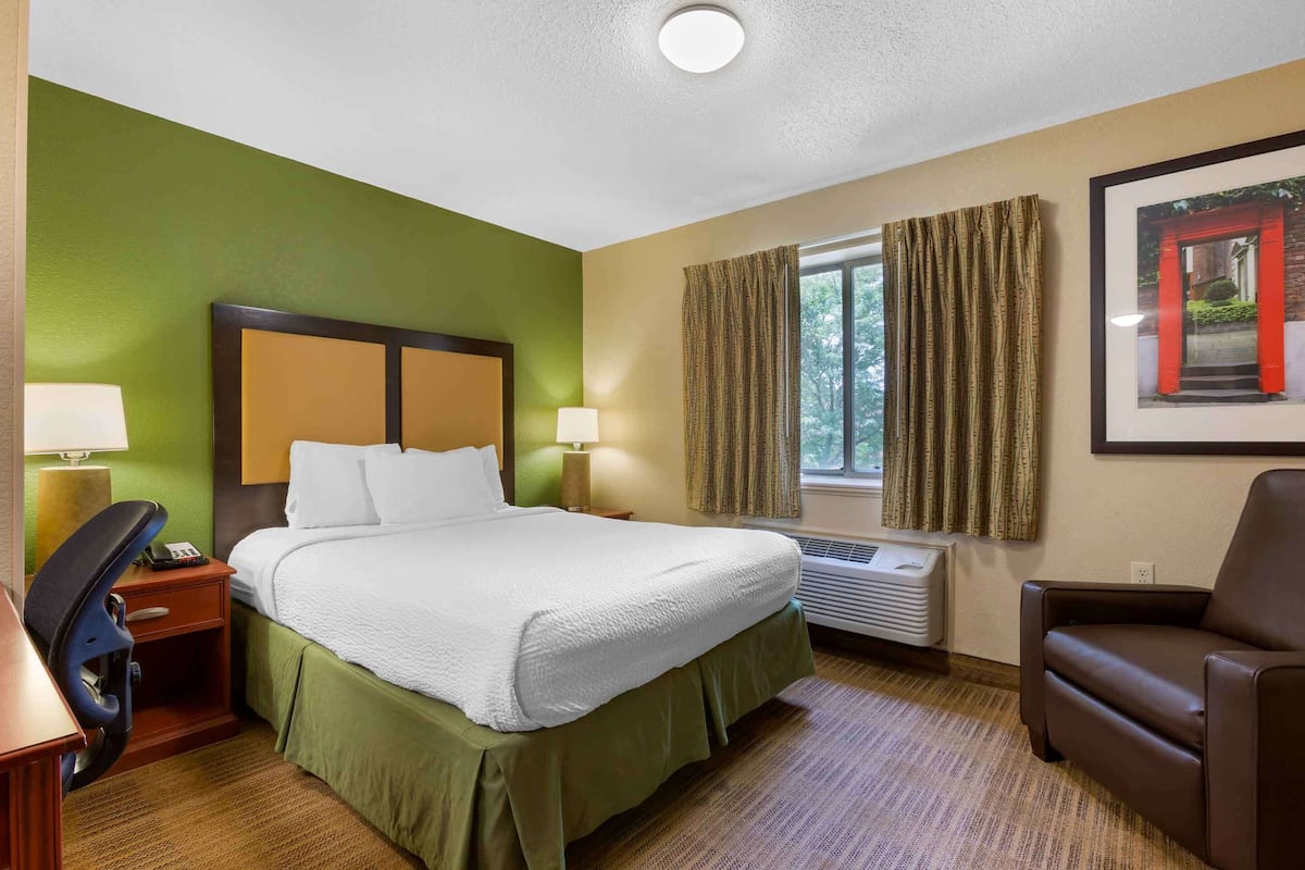 Ein Hotelzimmer mit einem Bett, einem Schreibtisch mit Stuhl, einer Couch, einem Fenster mit Vorhängen und einem gerahmten Bild an der Wand.