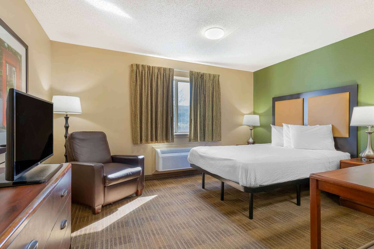 Habitación de hotel con cama, escritorio, silla, televisor y ventana con cortinas.