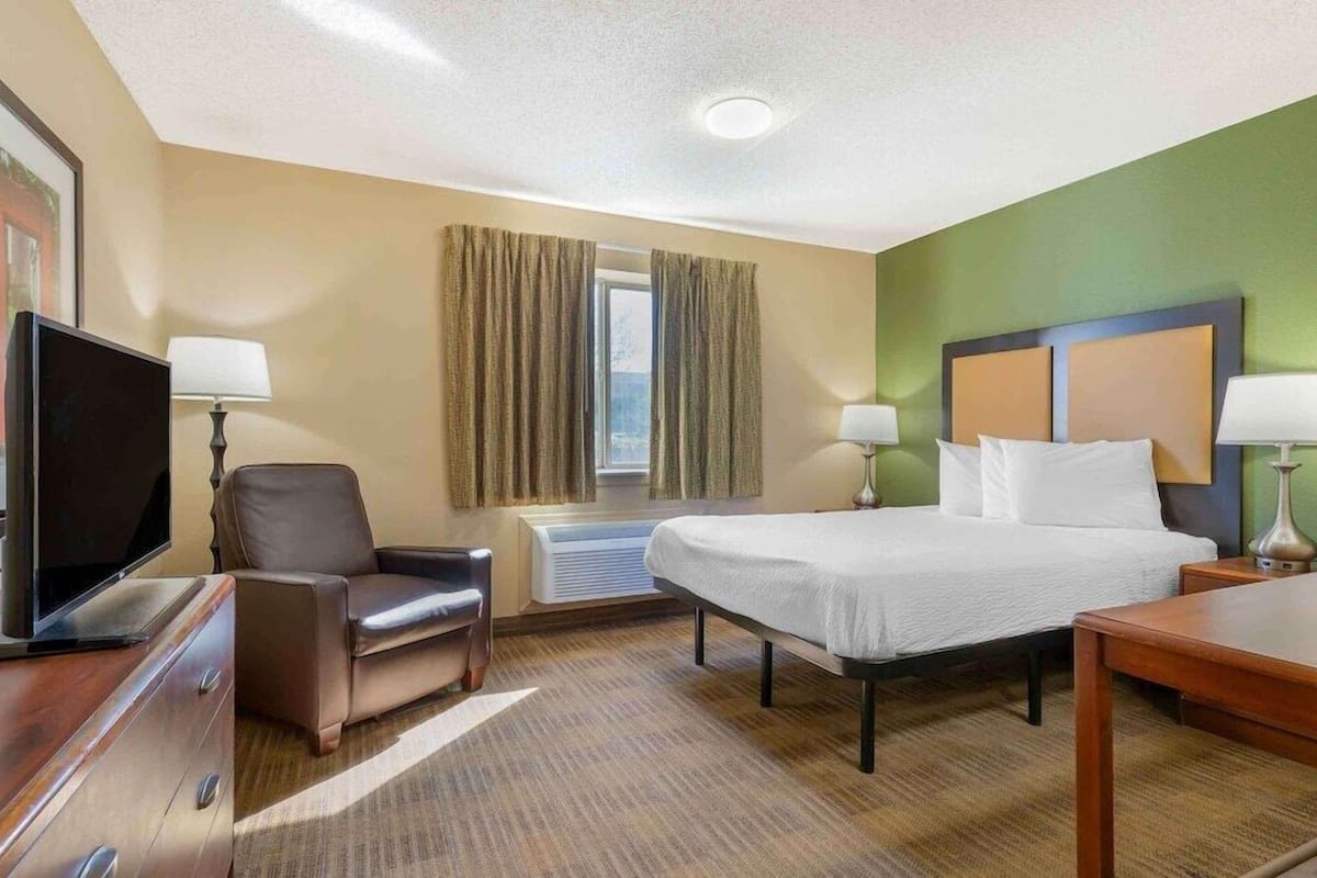 Una habitación de hotel con cama, escritorio, silla, televisor y ventana con cortinas.