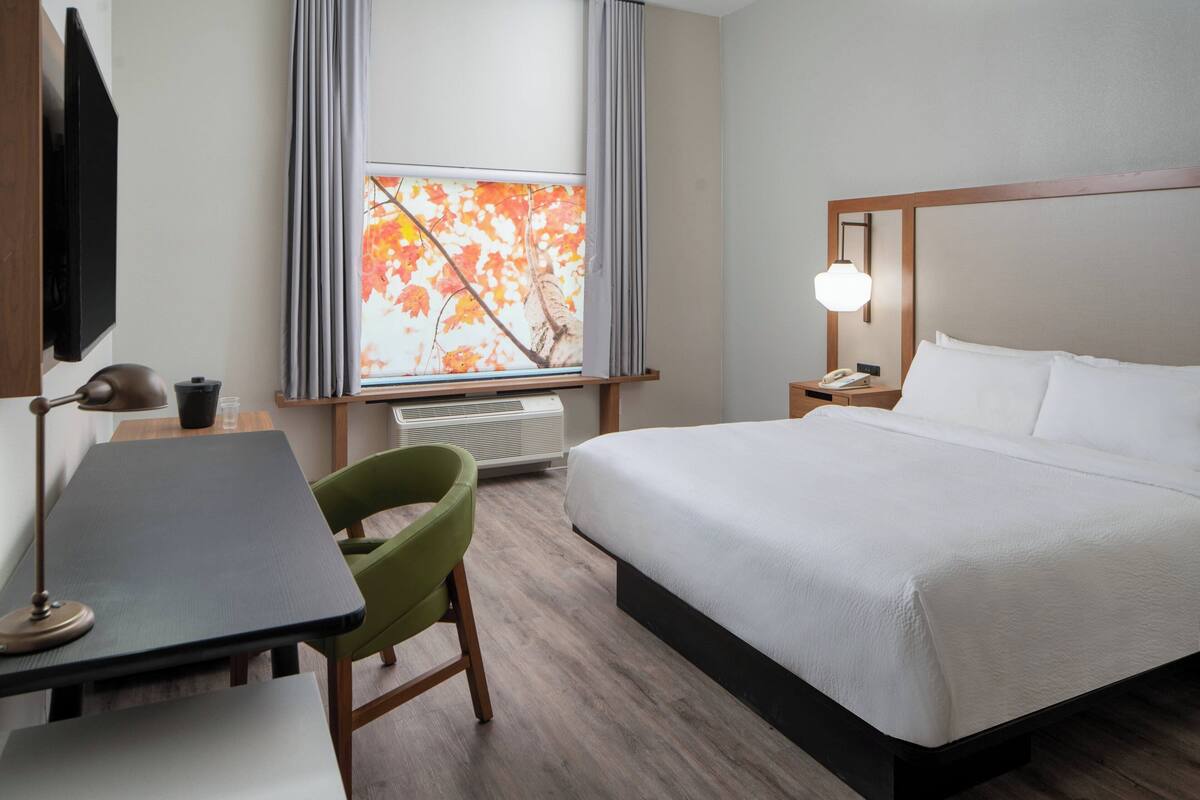 Une chambre d’hôtel comprenant un lit, un bureau, une chaise, une télévision, une fenêtre avec des rideaux et une lampe de chevet.