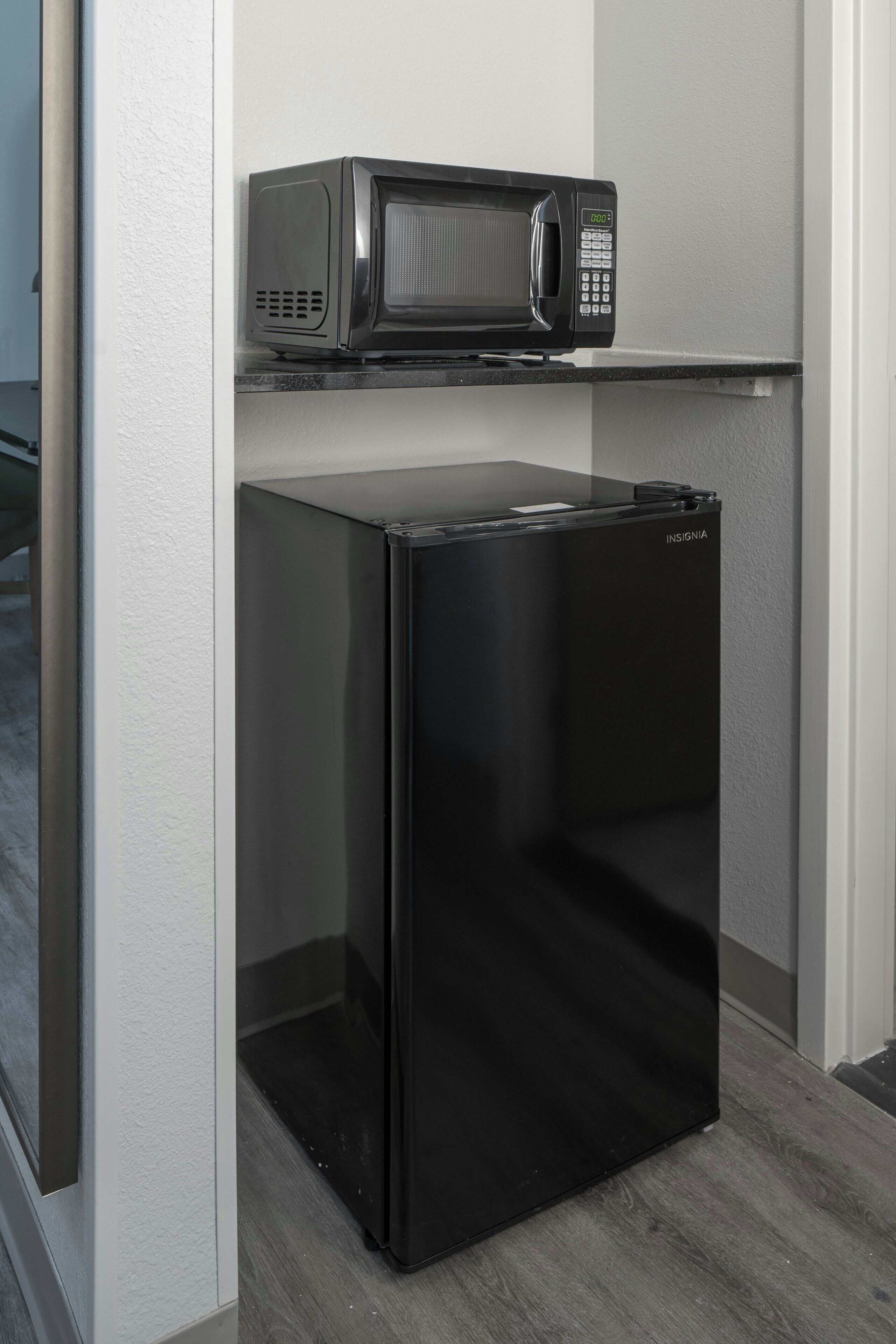Un refrigerador y microondas negros compactos en una pequeña cocina con pared blanca y piso de madera.