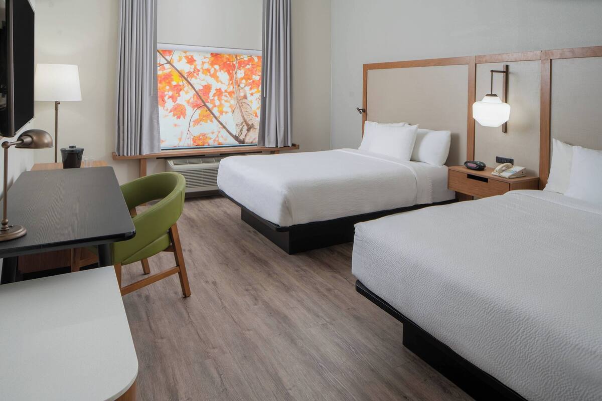 Une chambre d’hôtel avec deux lits, un bureau, une chaise, une lampe et une fenêtre avec des rideaux.