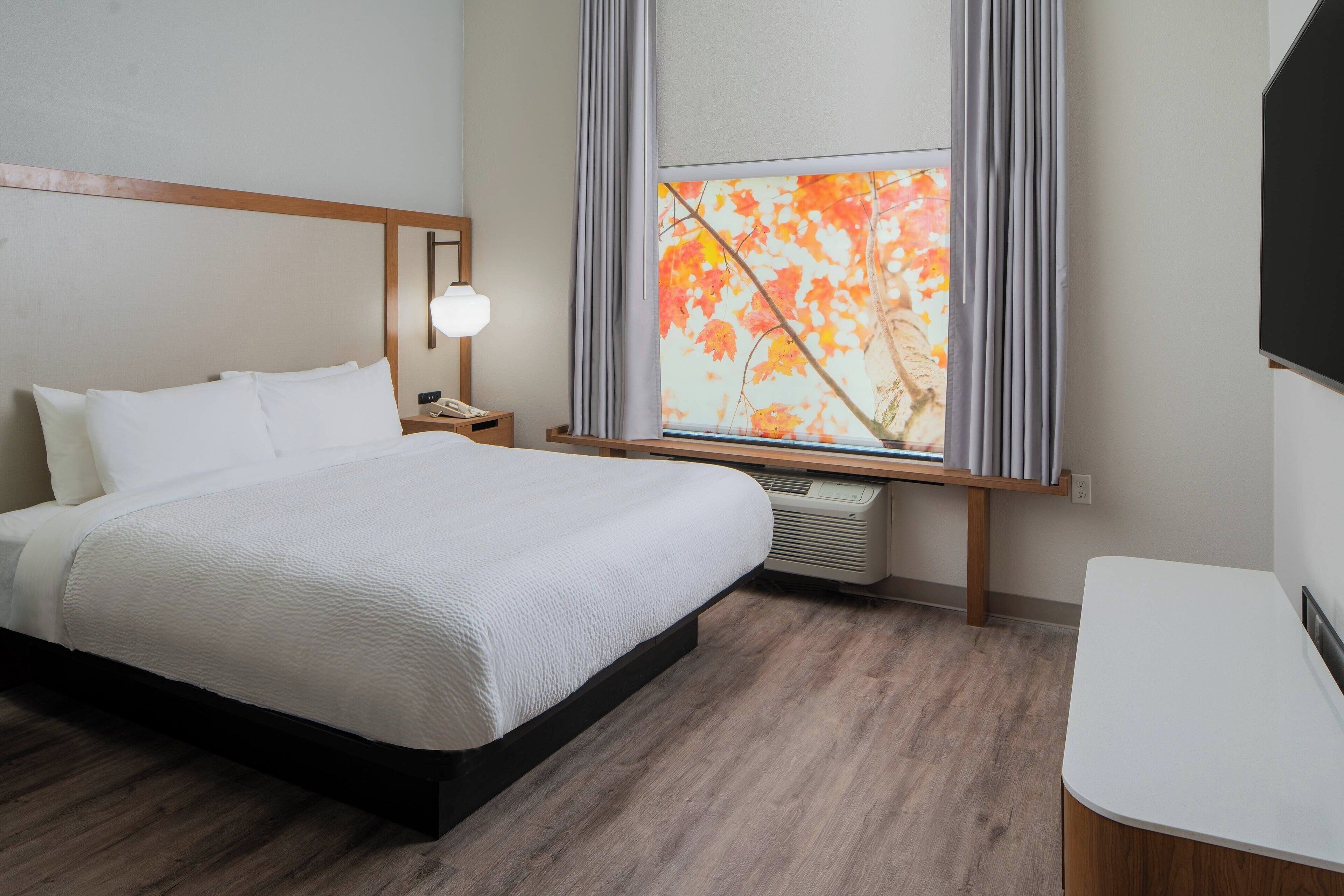A hotel room with a bed, bedside table, lamp, air conditioning unit, and a window with curtains.