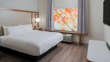 A hotel room with a bed, bedside table, lamp, air conditioning unit, and a window with curtains.