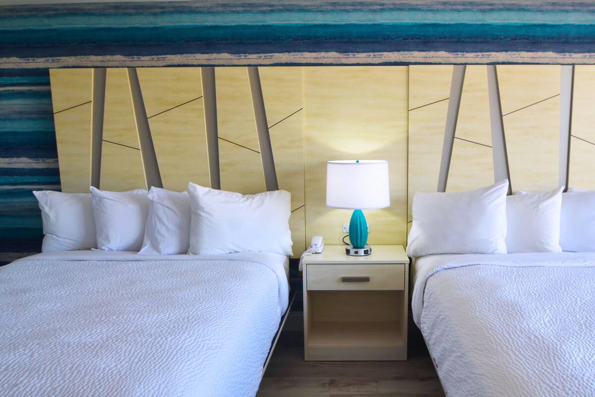 A hotel room with two beds, a nightstand with a lamp, and a wall with a blue and yellow pattern.