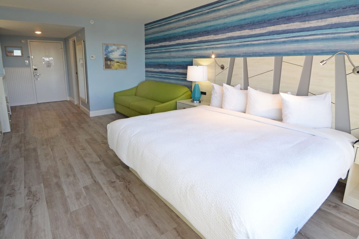 A hotel room with a large bed, a green chair, a desk with a TV, and a painting on the wall.