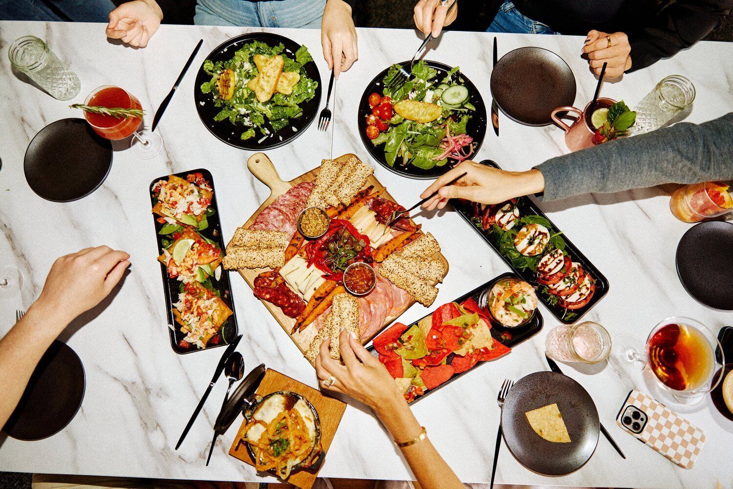 A group of people dining at a table with various dishes, including salads, meats, and cheeses, along with drinks and a smartphone.