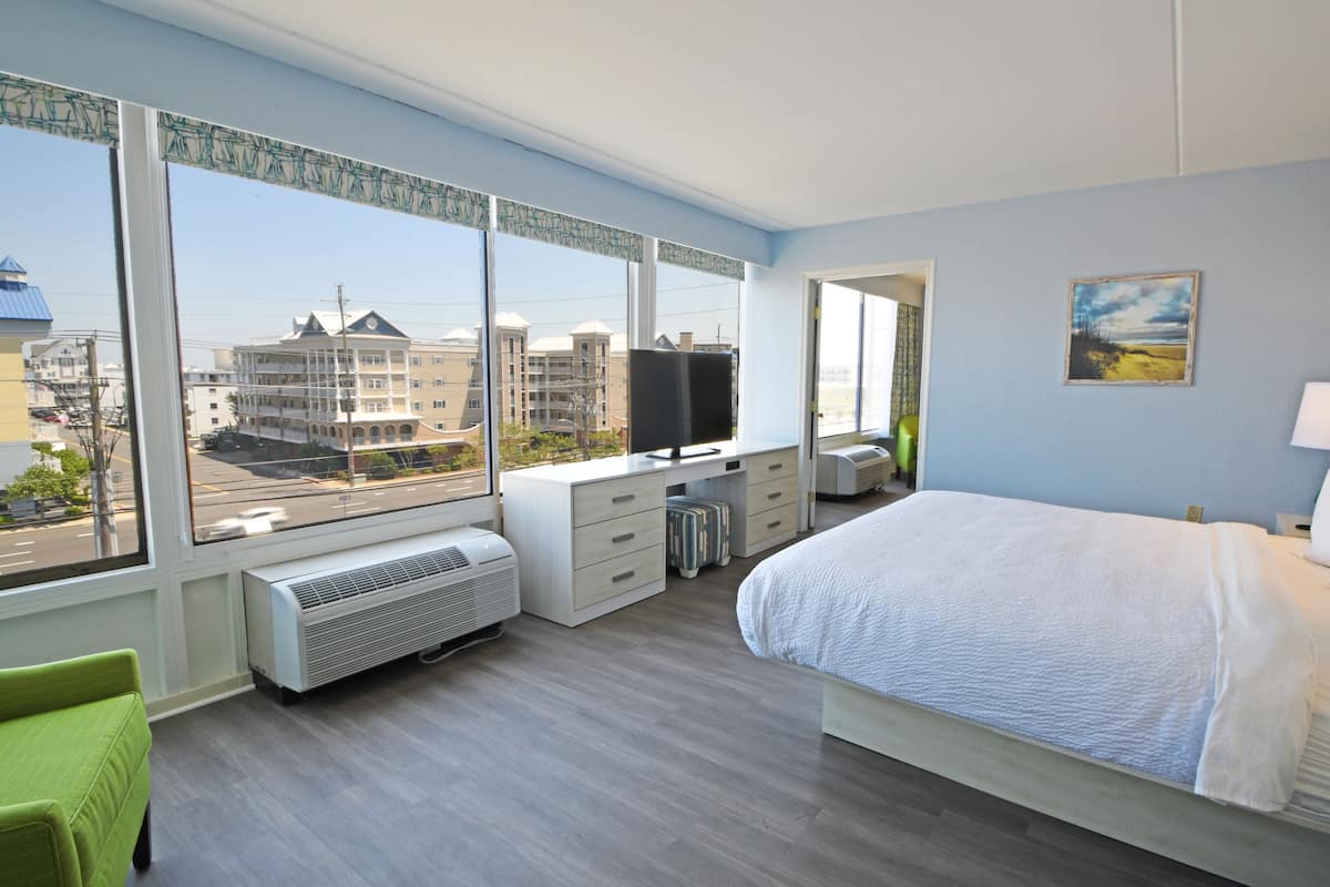 A modern hotel room with a large bed, a desk with a computer, a television, and a view of the city through large windows.