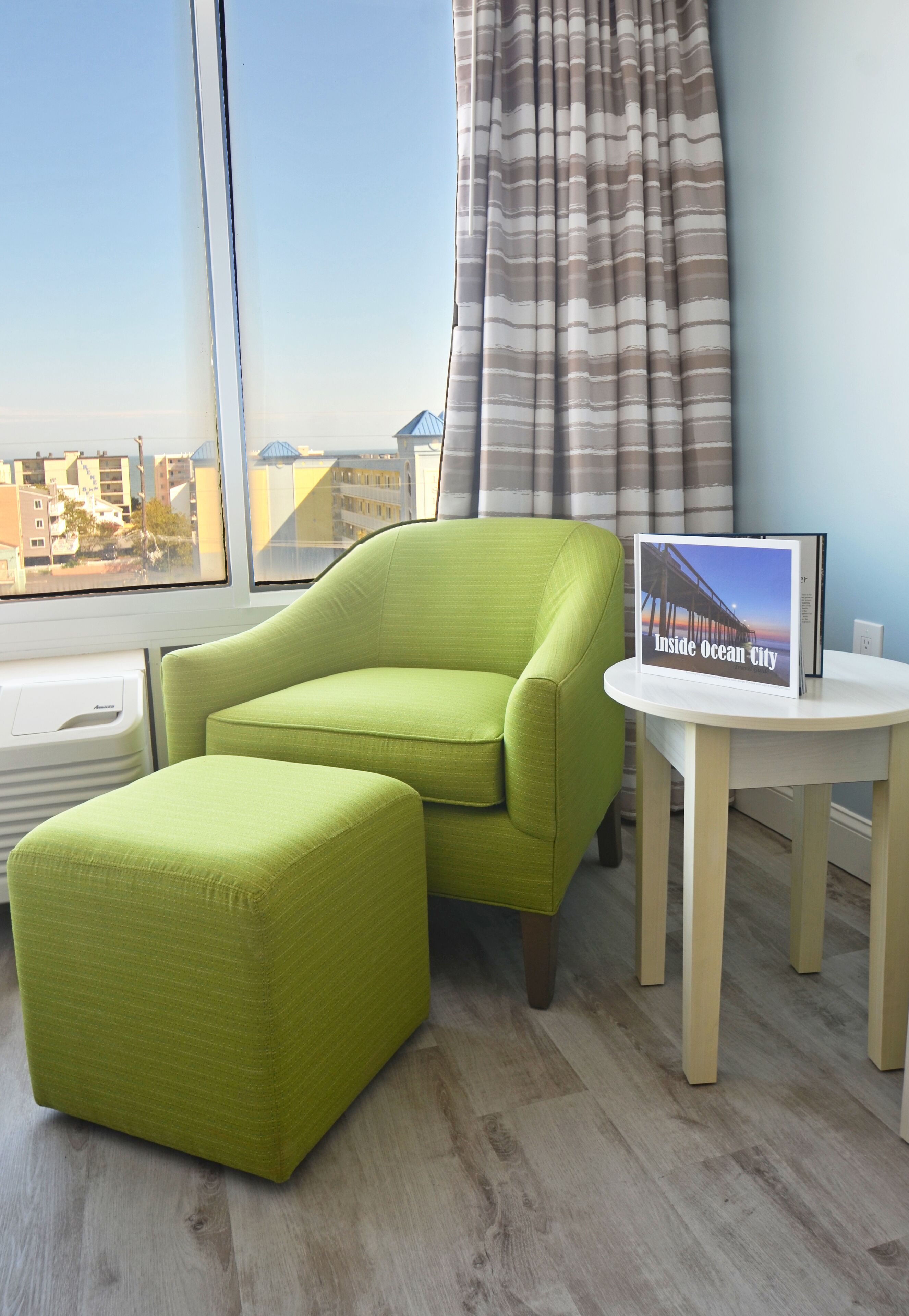 A room with a green armchair, a matching ottoman, a small table with a framed picture, and a window with a view of buildings.
