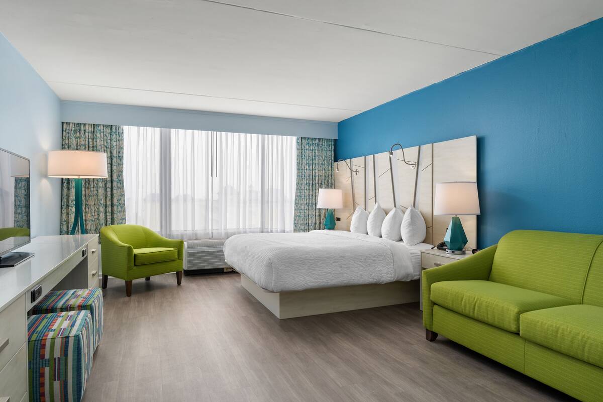 A hotel room with a bed, a green armchair, a desk with a computer, and a TV.