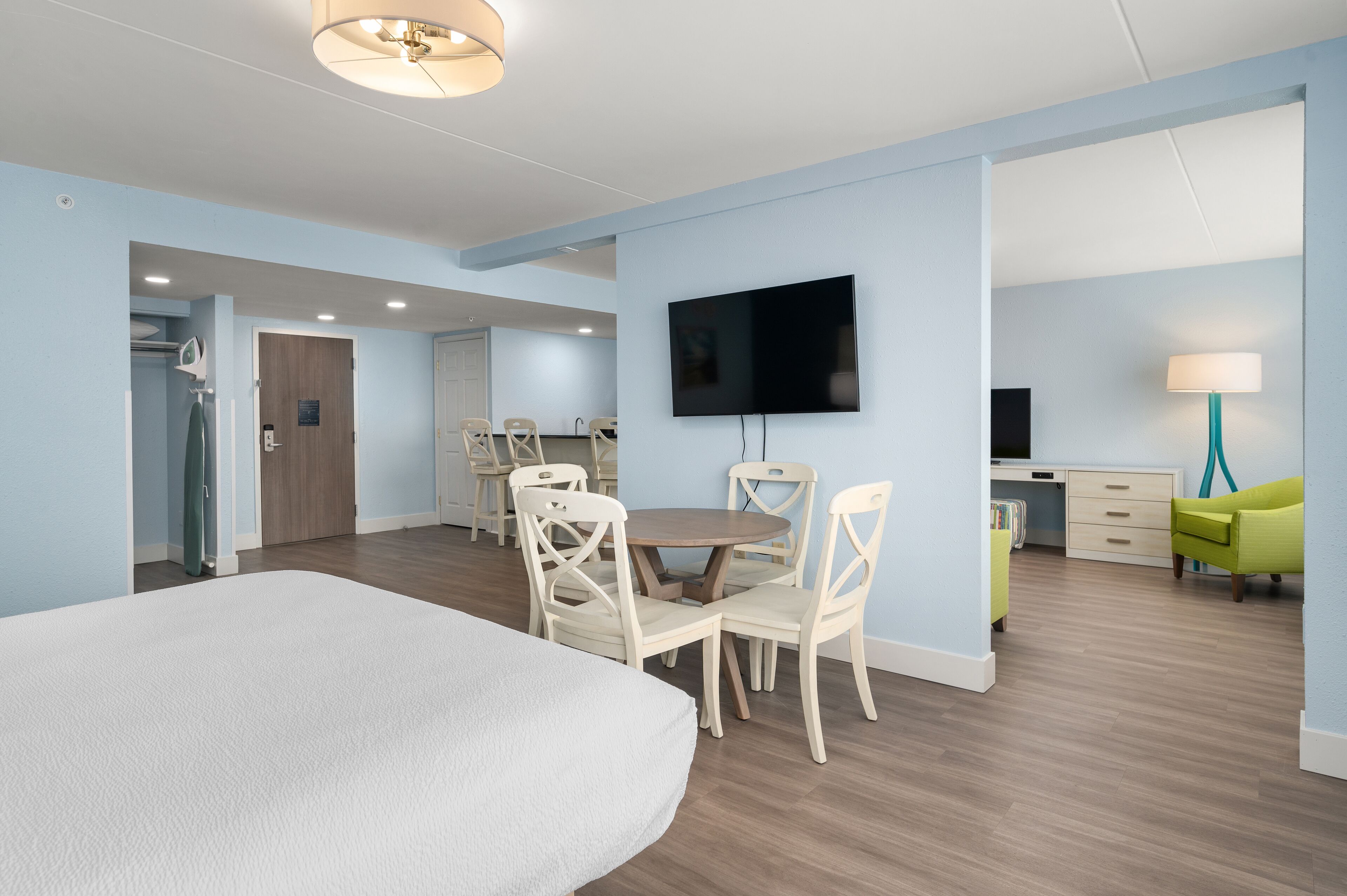 A modern hotel room with a large bed, a dining area with white chairs, a TV mounted on the wall, and a desk with a lamp.