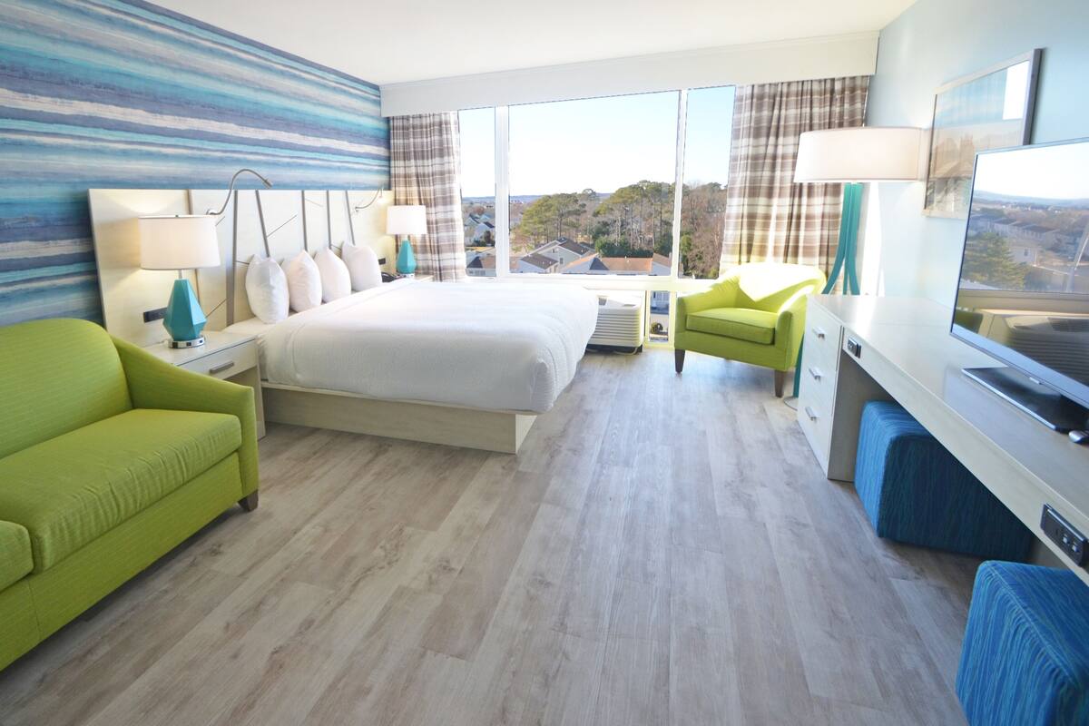A hotel room with a large bed, a green sofa, a desk with a TV, and a view of the outside.