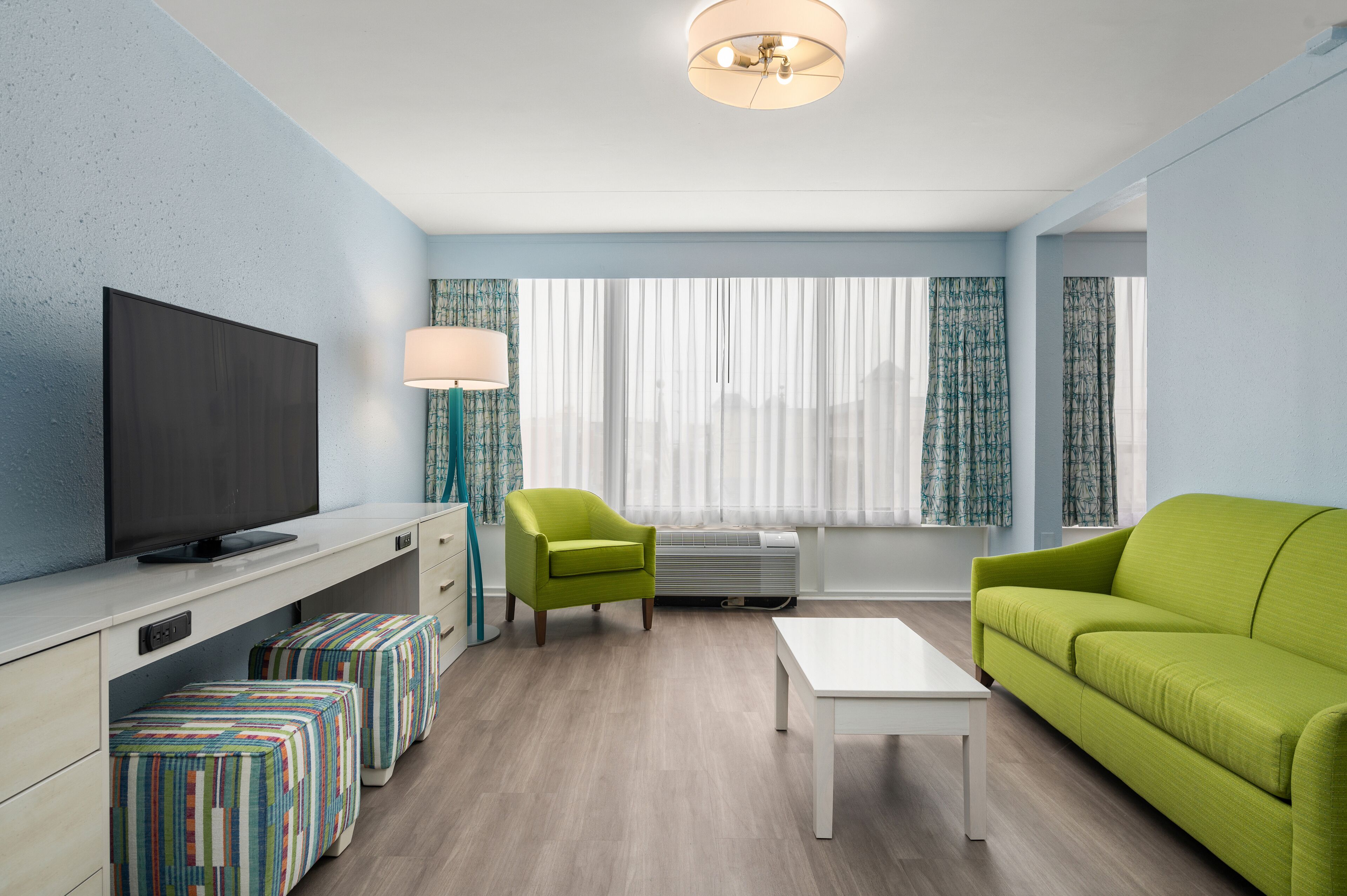 A modern living room with a flat-screen TV, green armchairs, a white coffee table, and striped ottomans.