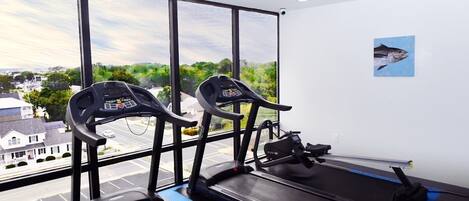 Une salle de sport équipée de deux tapis de course et d’un rameur, avec de grandes fenêtres donnant sur un quartier résidentiel.