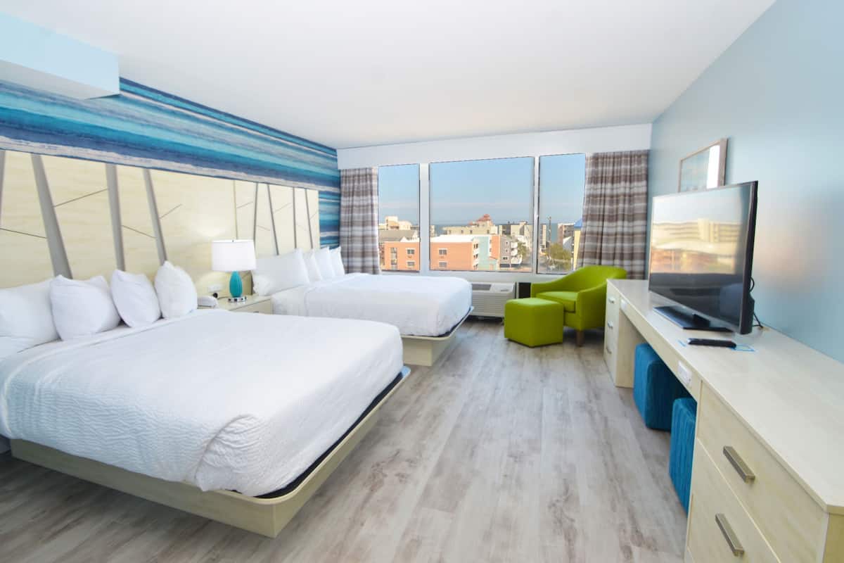 A hotel room with two beds, a desk with a TV, and a view of the city through large windows.