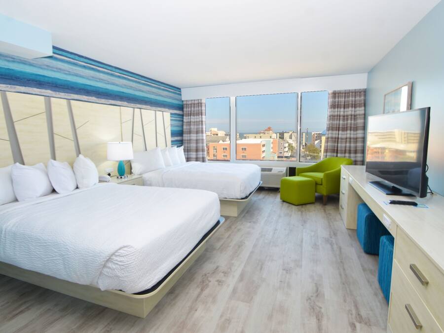 A hotel room with two beds, a desk with a TV, and a view of the city through large windows.