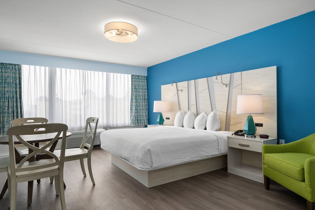 A hotel room with a bed, two chairs, a desk, a nightstand with lamps, and a view of the city through large windows.