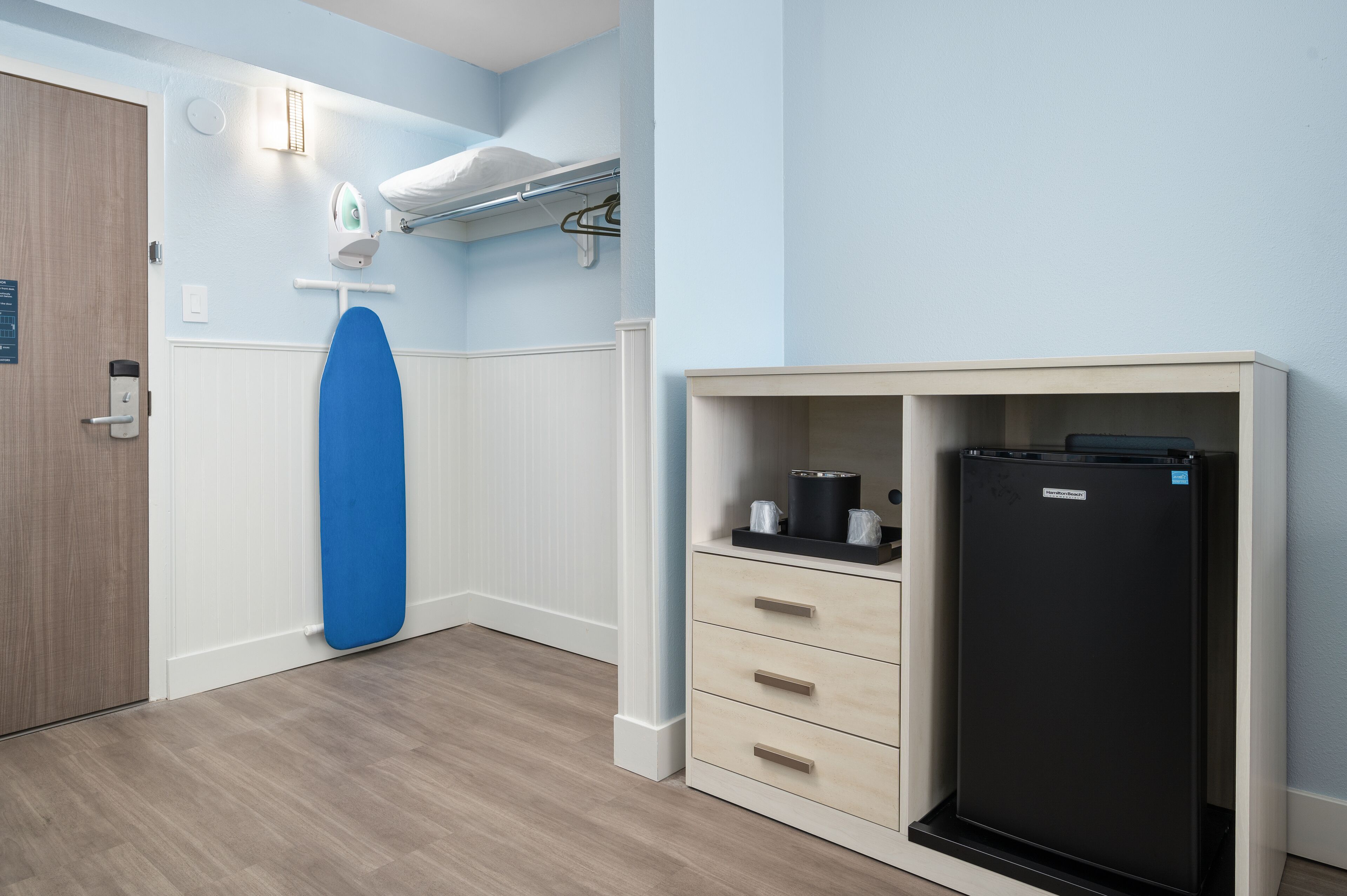 A room with a white door, a blue ironing board, a black refrigerator, and a wooden cabinet with drawers.