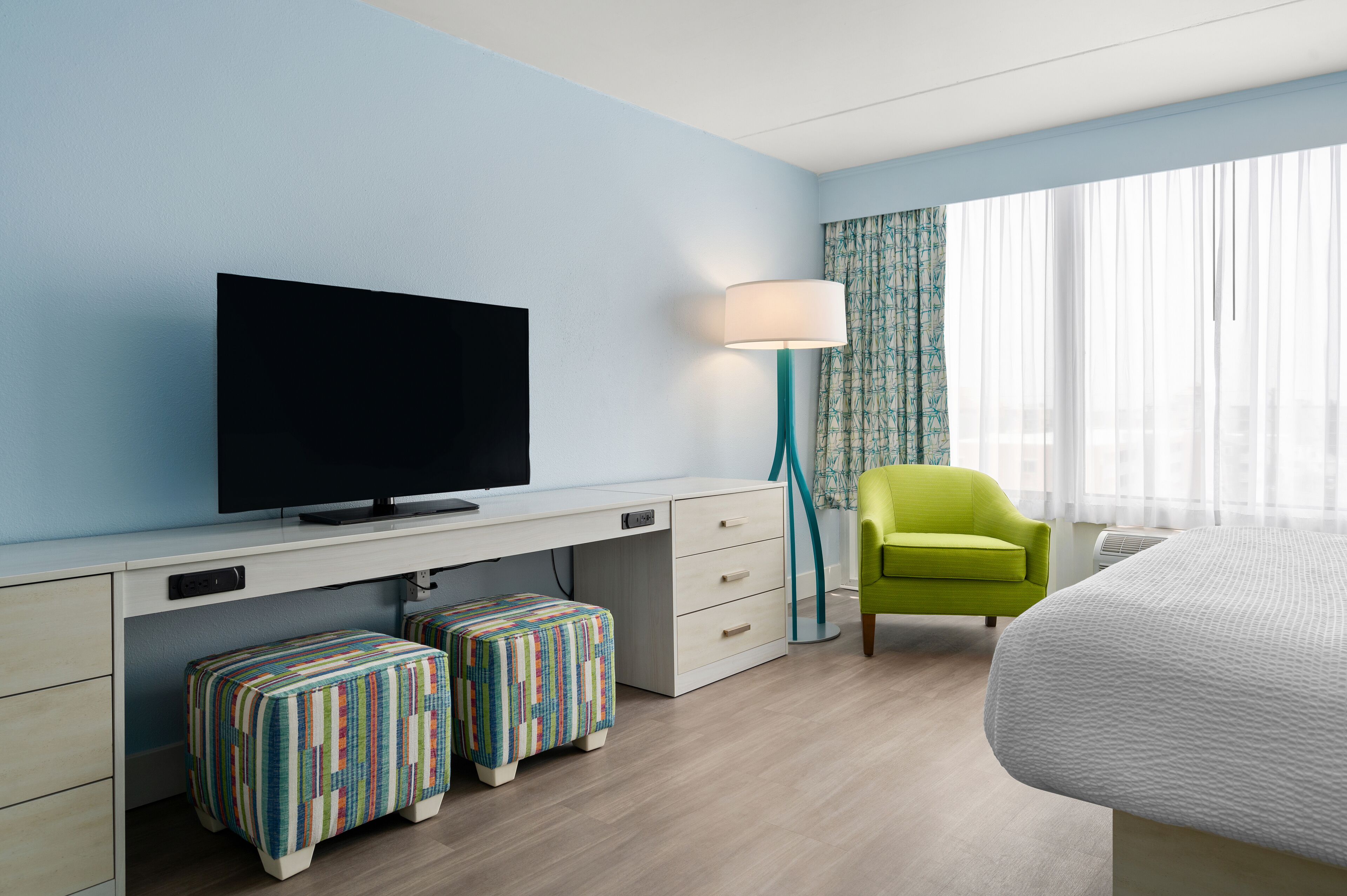 A hotel room with a flat-screen TV, a desk with drawers, a colorful ottoman, a green chair, and a bed.