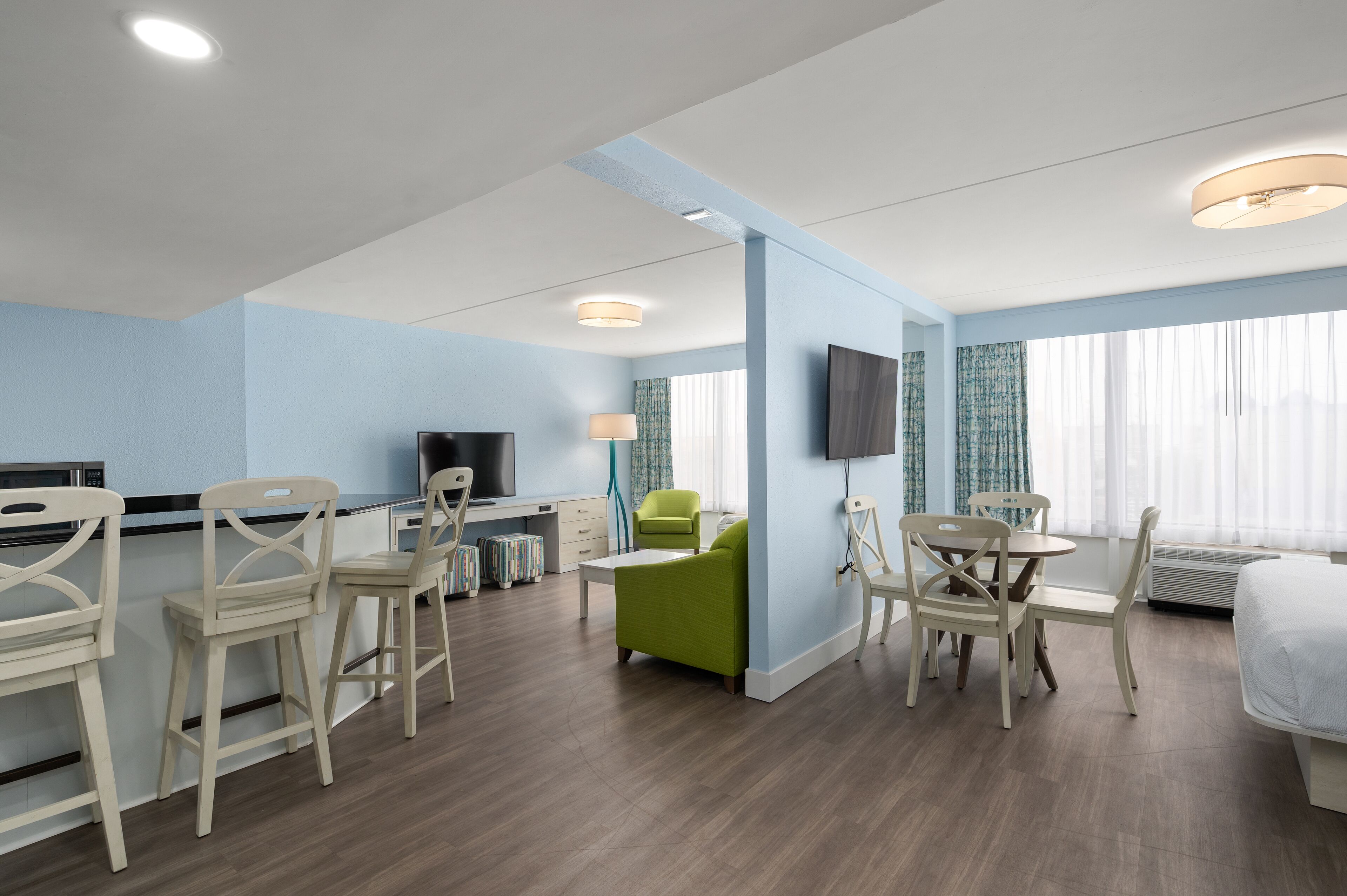 Quarto de hotel moderno com cozinha compacta, televisão, poltrona verde e uma cama.