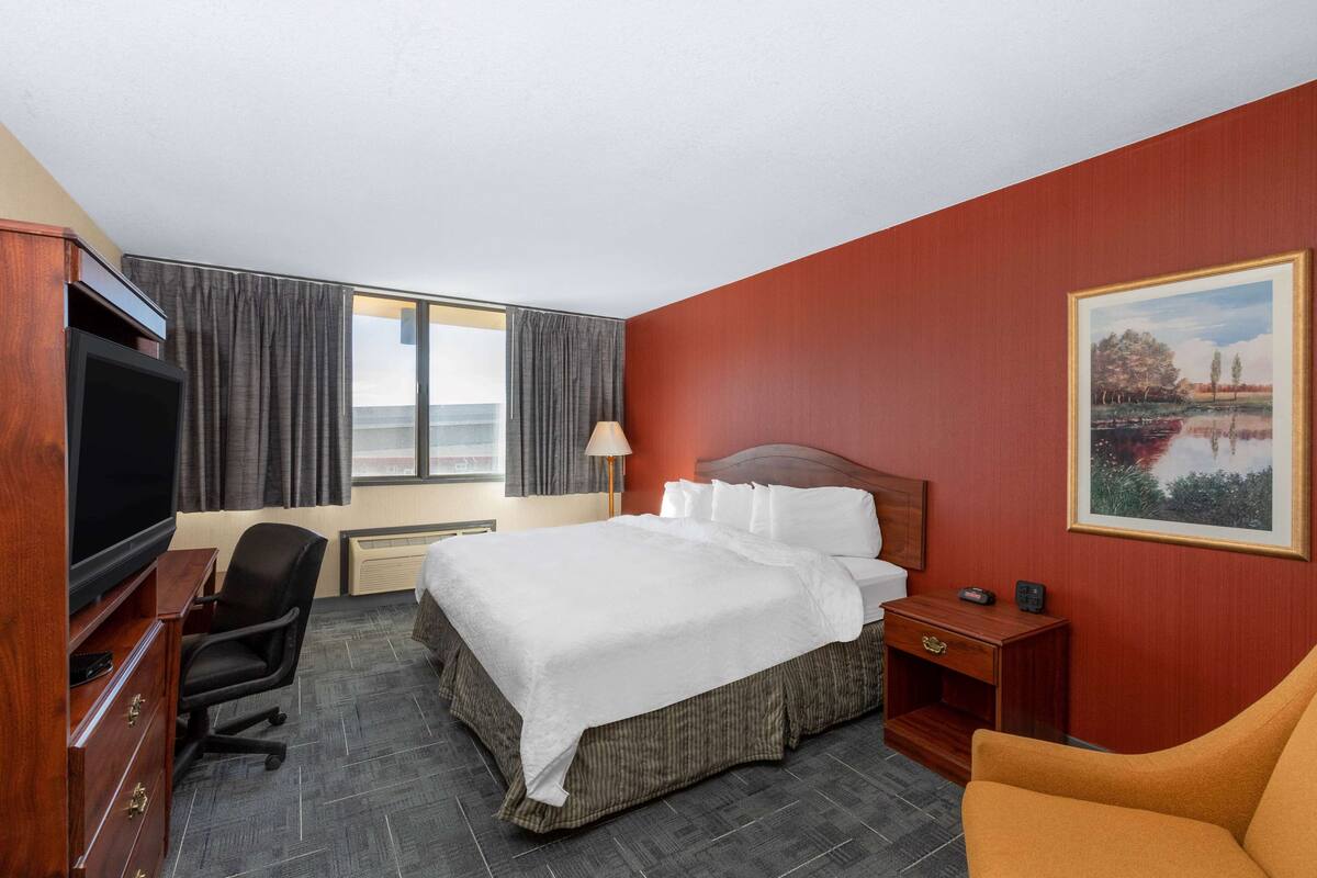 A hotel room with a bed, a desk with a television, a chair, a nightstand, and a painting on the wall.