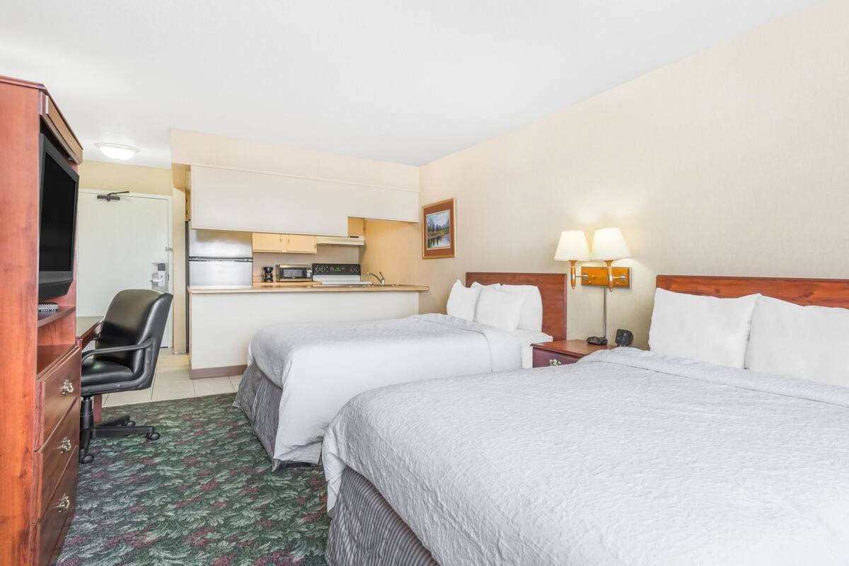 A hotel room with two beds, a desk with a chair, a kitchenette, and a TV.