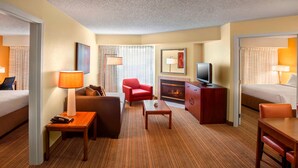 Habitación de hotel con una cama, un sofá, un sillón rojo, una mesita, un televisor, una chimenea y un escritorio con una lámpara.