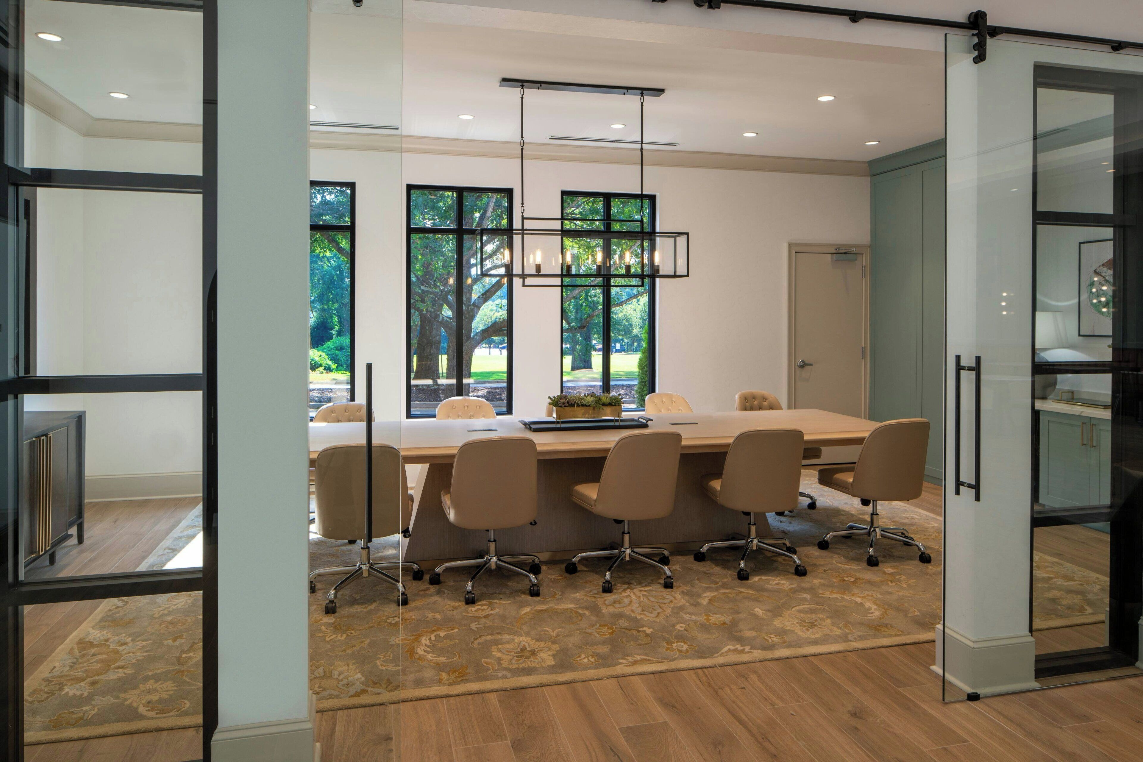 A conference room with a long table, four chairs, a chandelier, and large windows with a view of trees.