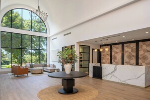 A spacious lobby with a round table, sofas, a reception desk, and large windows offering a view of trees.