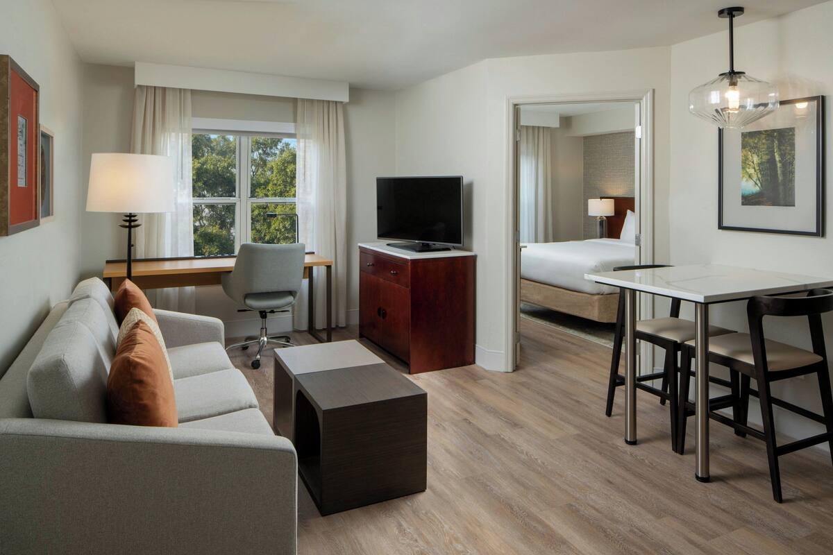A modern hotel room with a sofa, a TV, a desk with chairs, a bed, and a window with curtains.