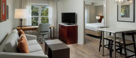 A modern hotel room with a sofa, a TV, a desk with chairs, a bed, and a window with curtains.