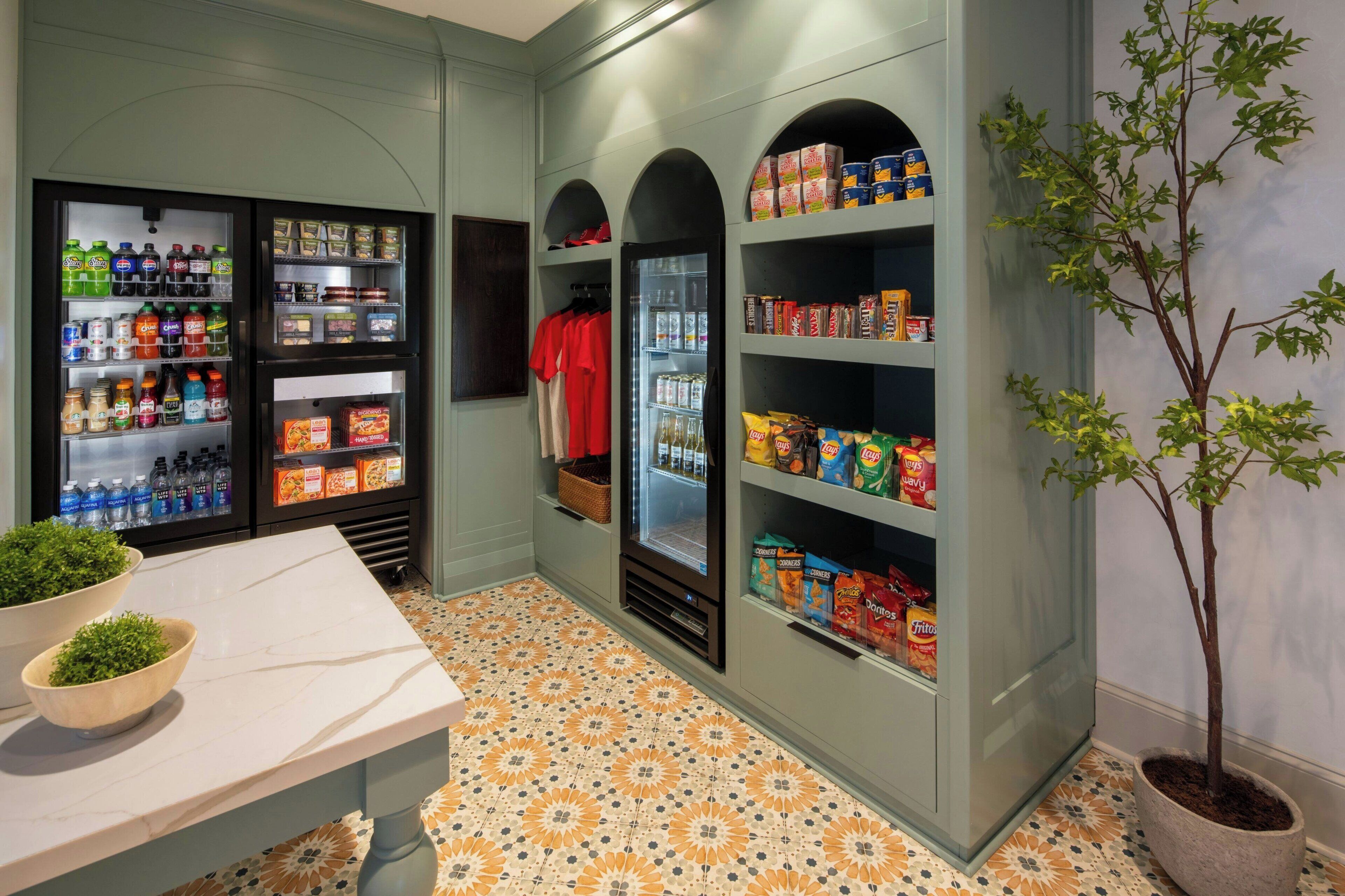 A kitchenette with a marble countertop, a refrigerator stocked with beverages and snacks, and a potted plant.